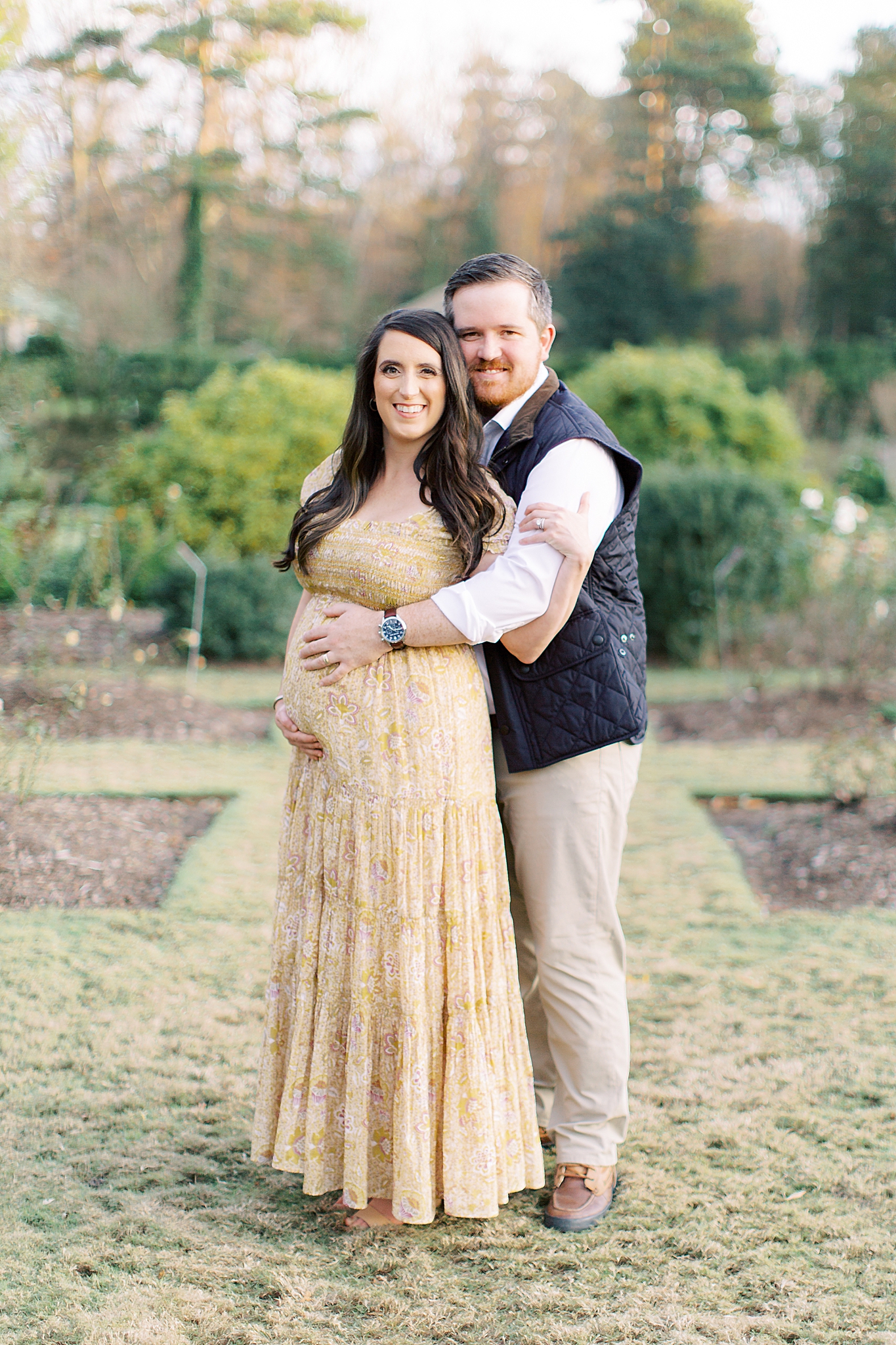 husband and wife pose in gardens at Reynolda Village during maternity photos