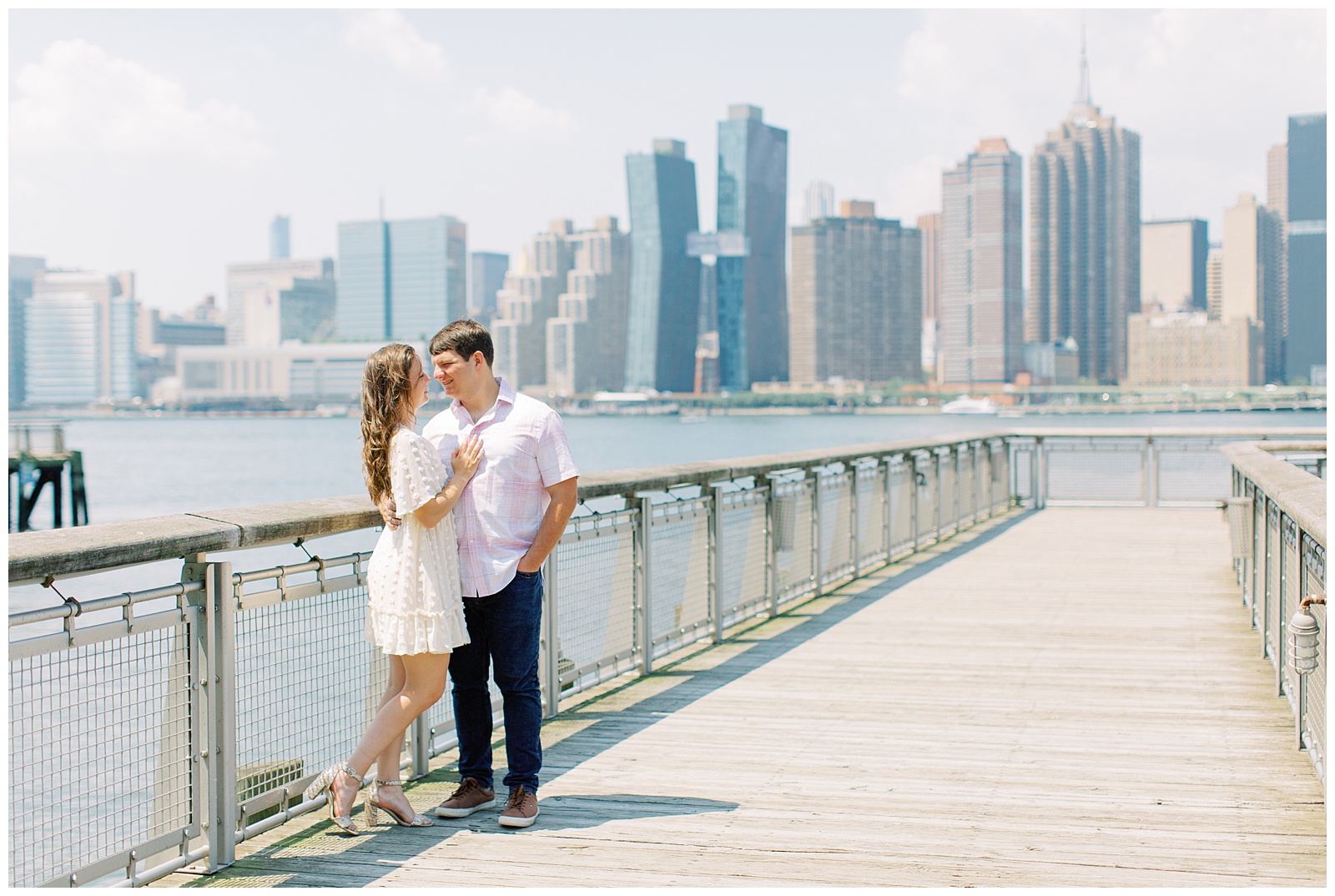 couple stands on pier with Manhattan skyline behind them during engagement photos 