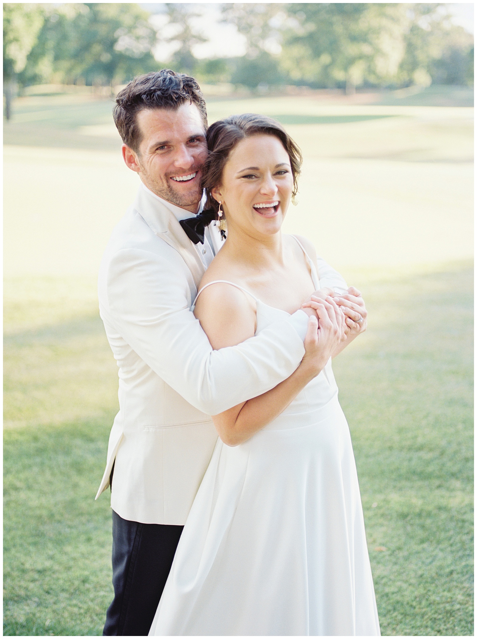 groom hugs bride from behind in white suit jacket at Quail Hollow Country Club
