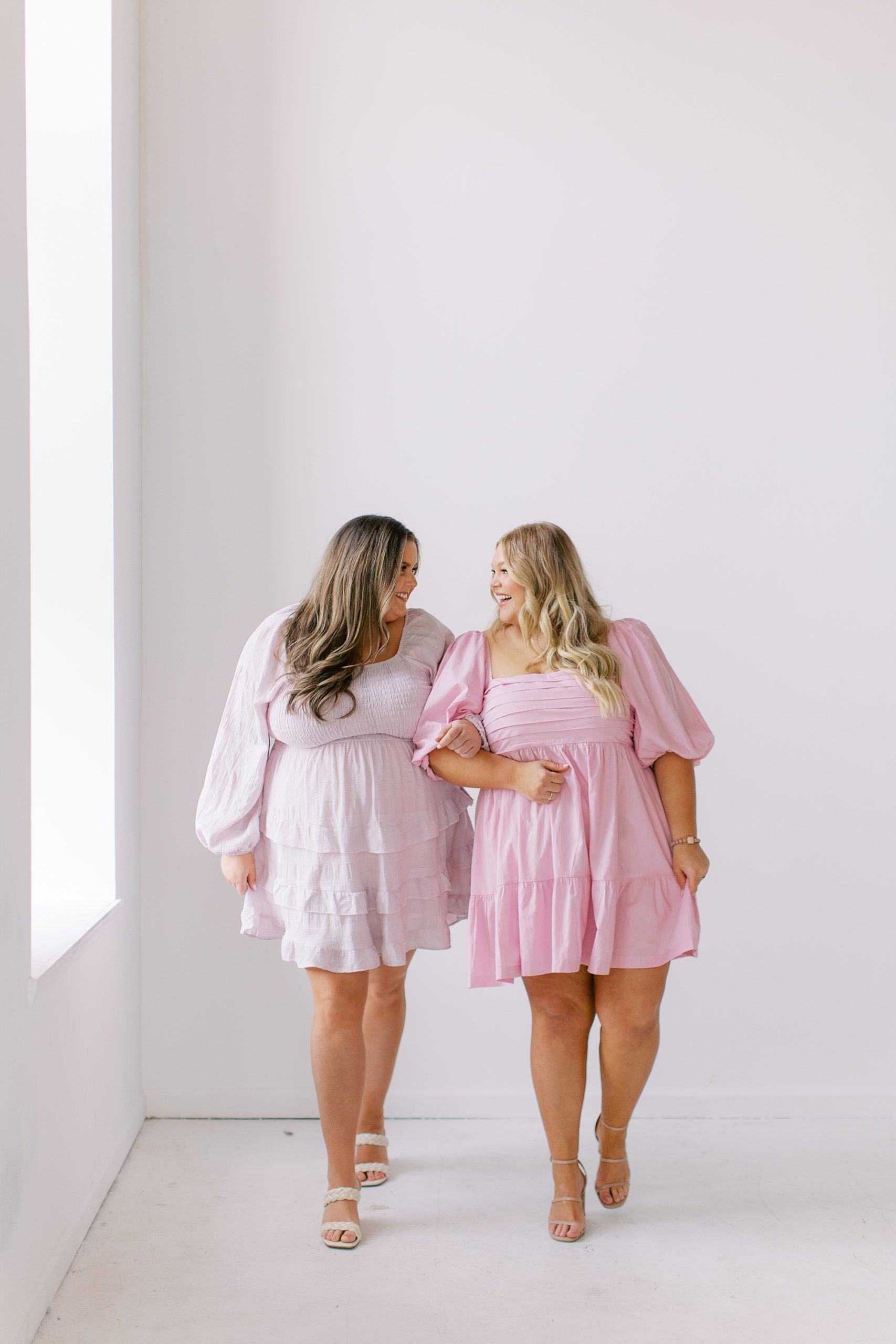 women link arms in pink dresses and walk in white studio