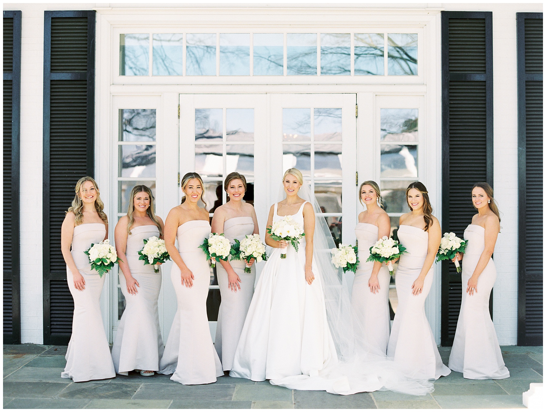 bride laughs with bridesmaids holding all-white bouquets outside double doors at Quail Hollow Club