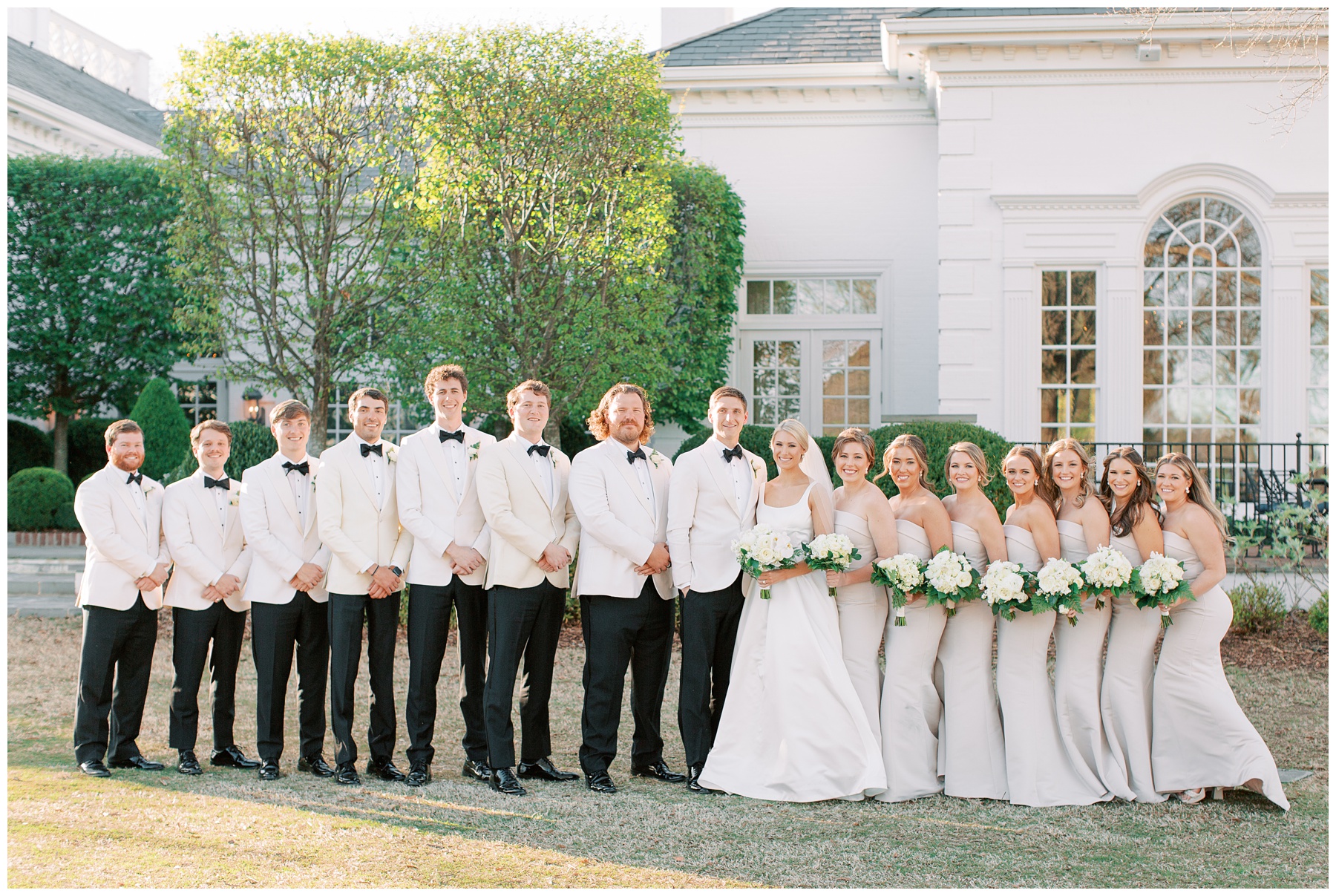 bride and groom stand with wedding party in white tux jackets and grey bridesmaid dresses at Quail Hollow Club
