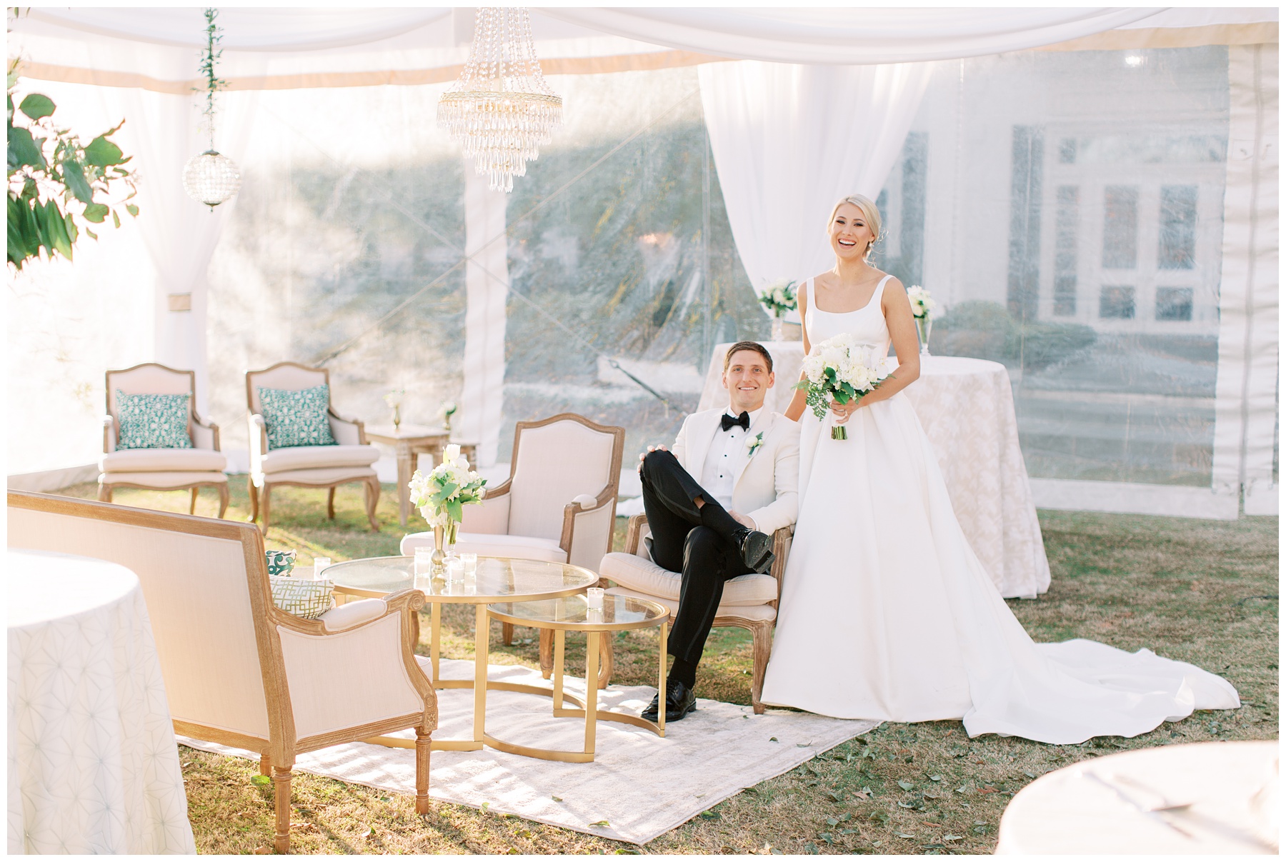 groom in white tux jacket sits in chair in tent with bride standing next to him