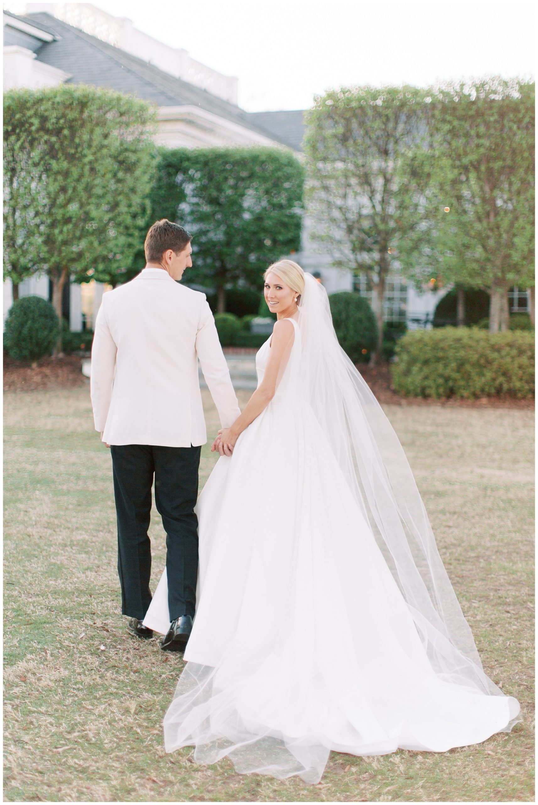 newlyweds hold hands walking towards Quail Hollow Club while bride looks over shoulder