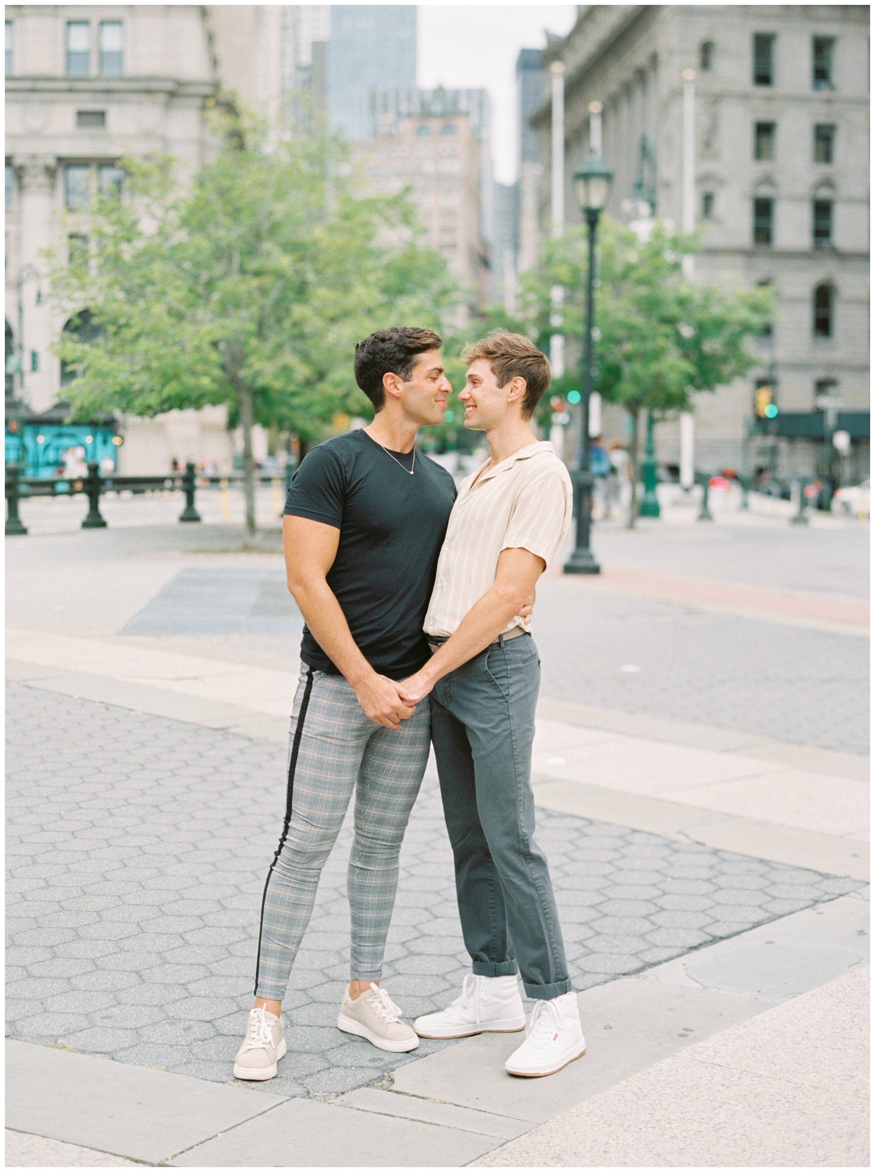 two men hug leaning heads together to kiss holding hands in Foley Square during New York City engagement session