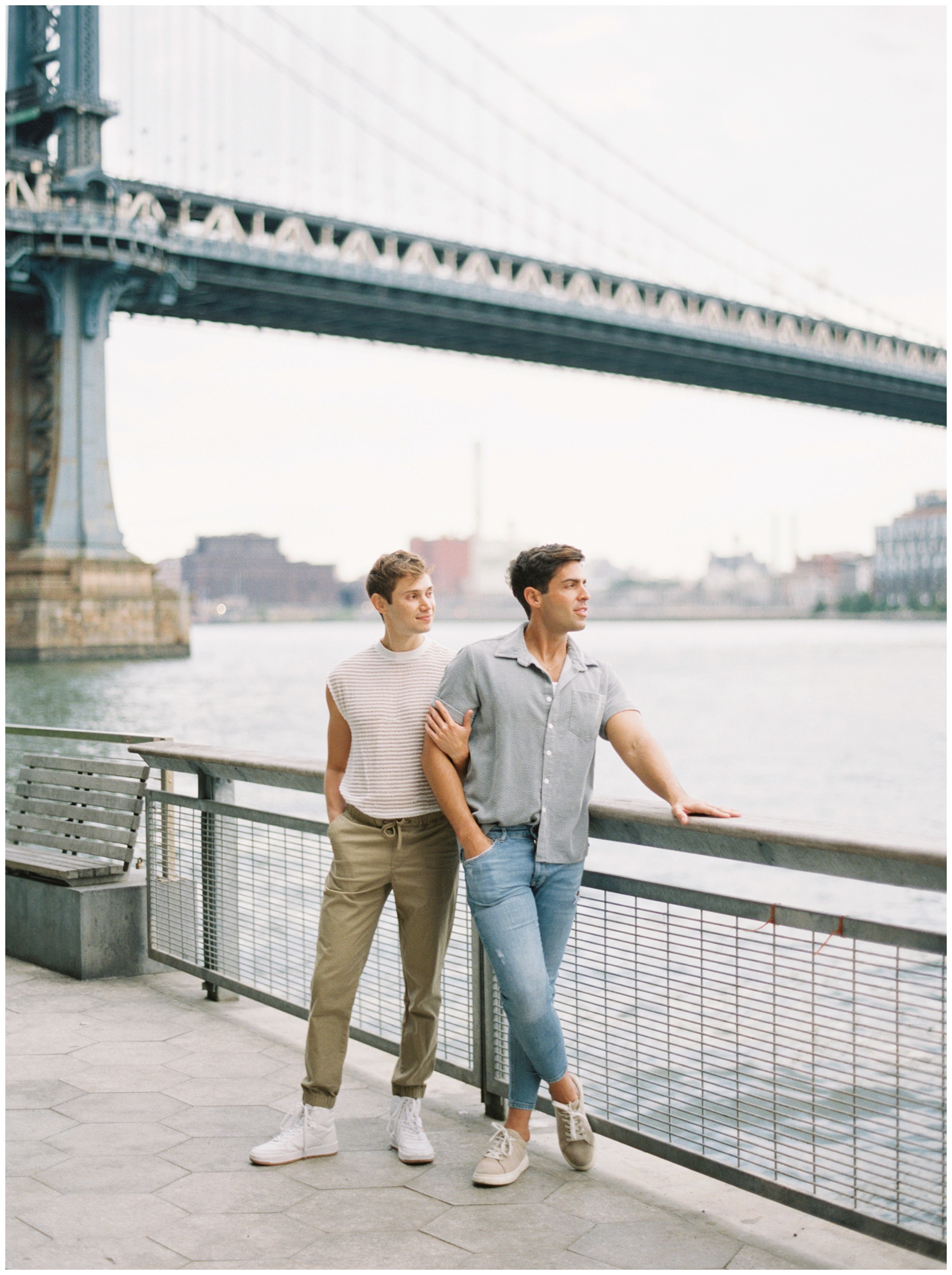 engaged men lean against railing near Two Bridges in New York City during engagement session 