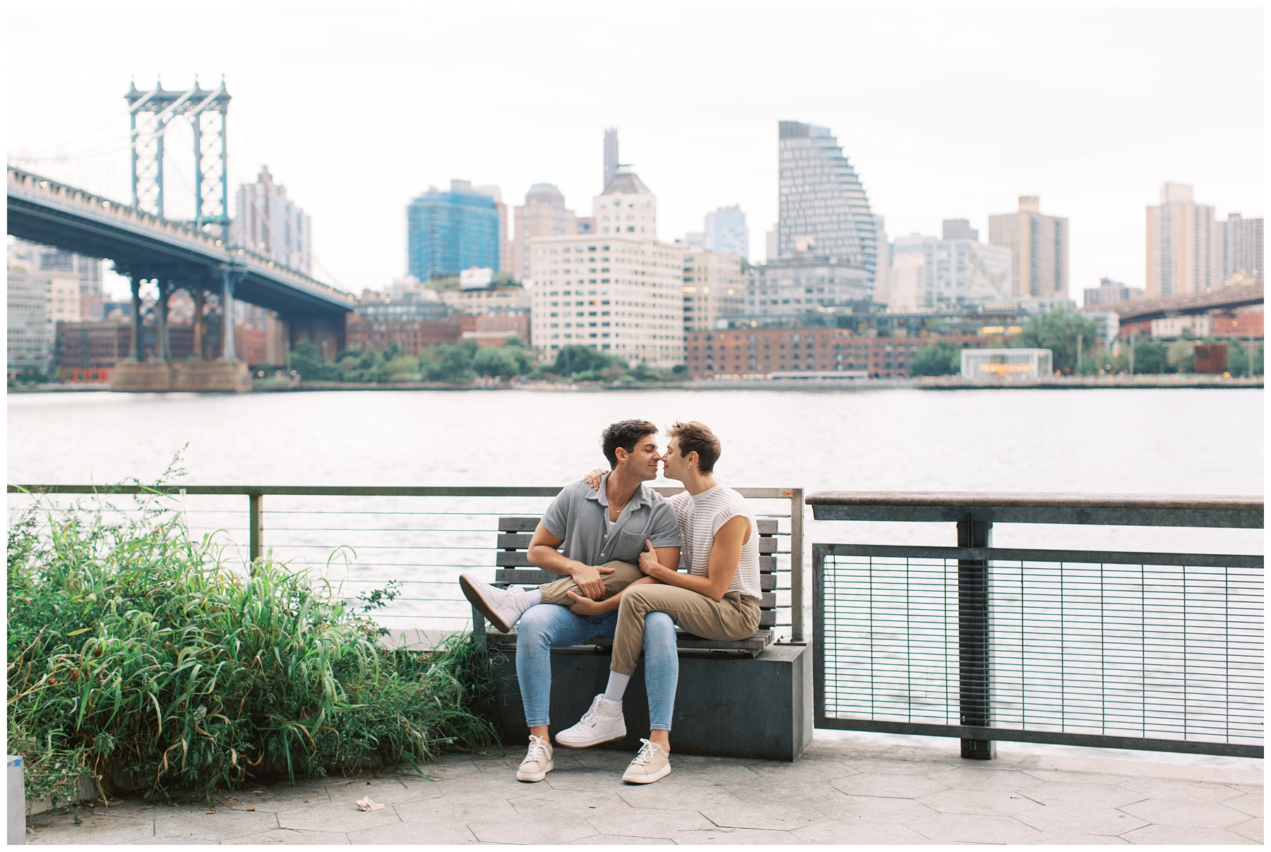 engaged couple sits together on park bench in front of NYC skyline in waterfront park 