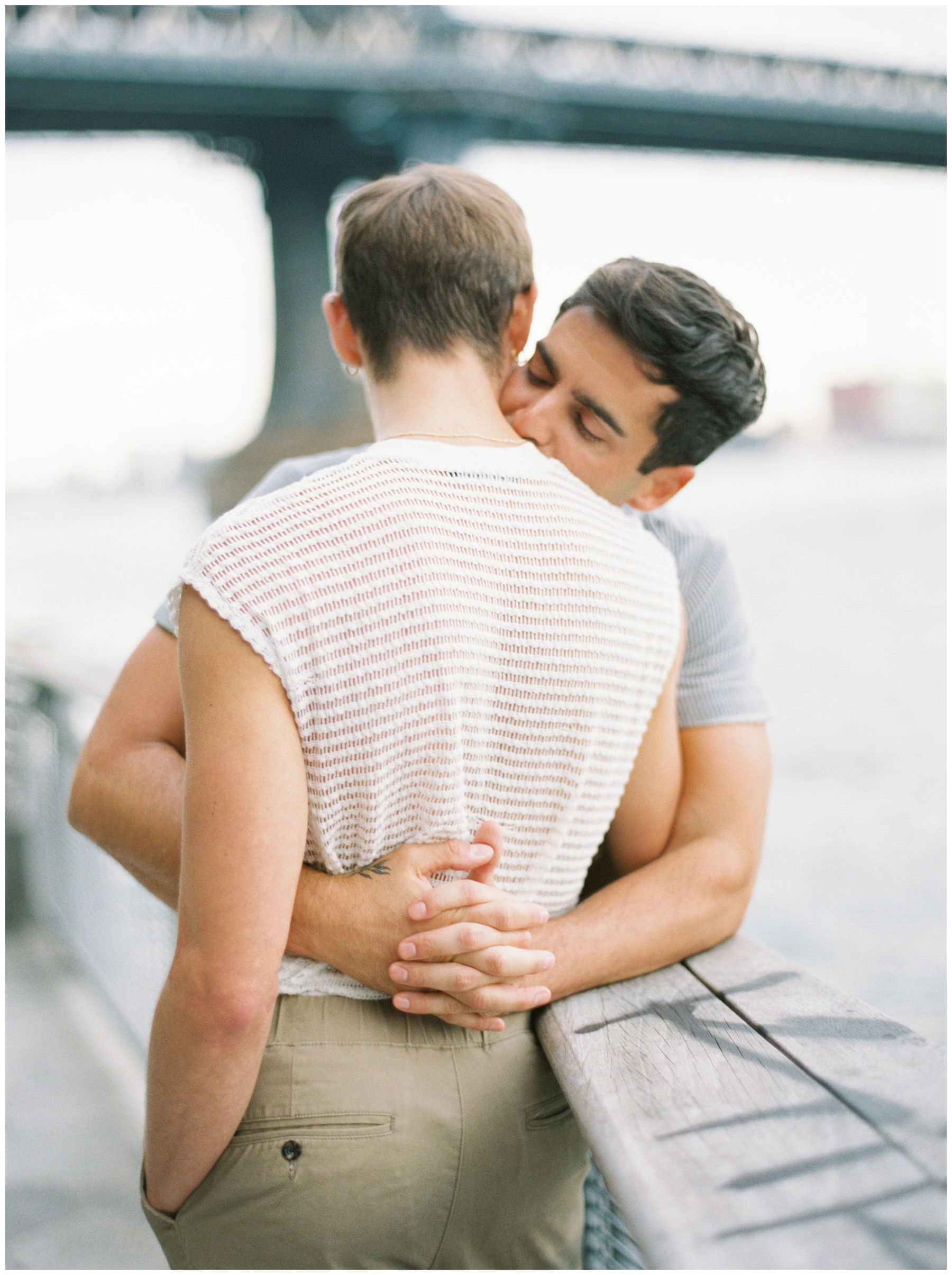 man in grey shirt hugs boyfriend around waist leaning to kiss his neck during NYC couples portraits 