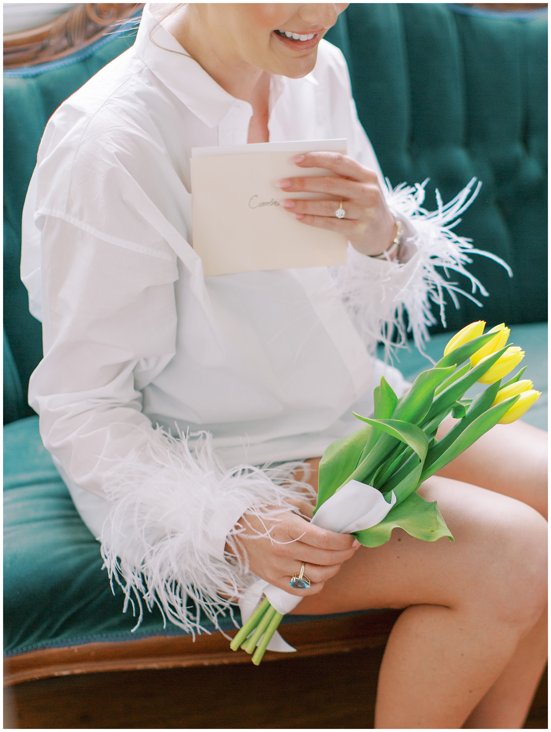 bride sits on teal couch holding yellow tulips and card from groom in shirt with feather cuffs