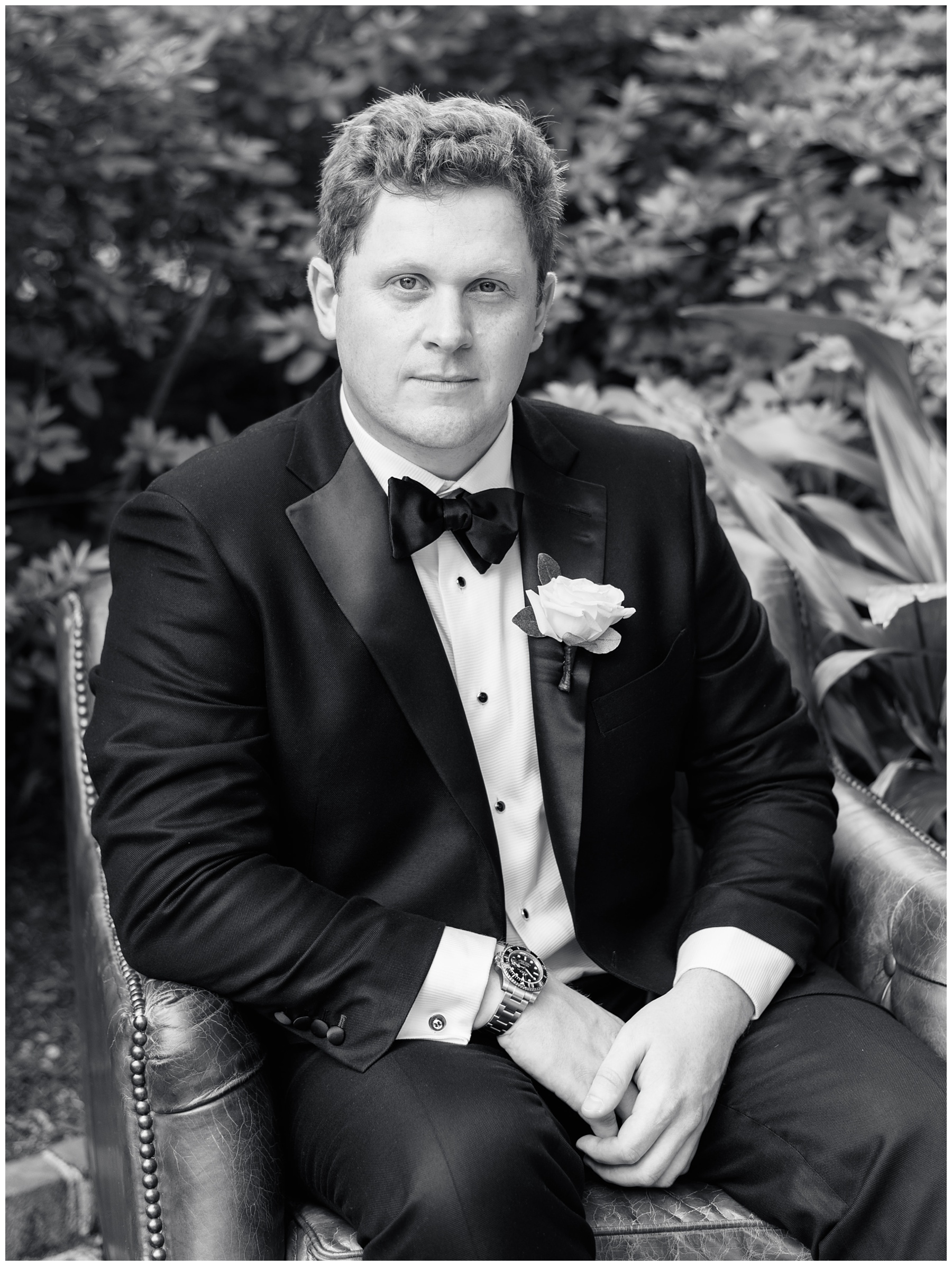 groom sits on leather couch outside in Charleston SC in black tux jacket 