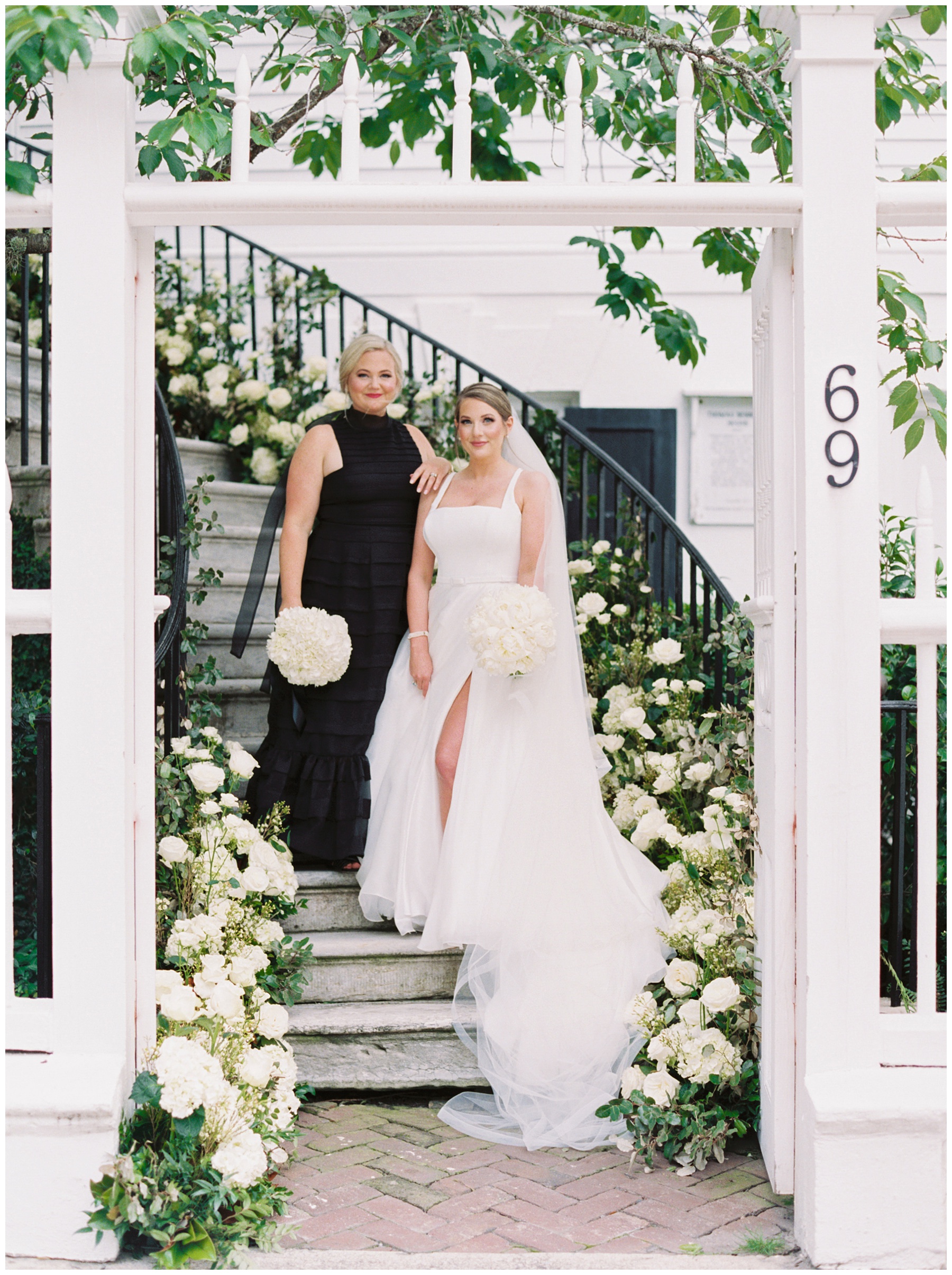 maid of honor stand on step above bride leaning on her on staircase at the Governor Thomas Bennett House covered in ivory roses and greenery