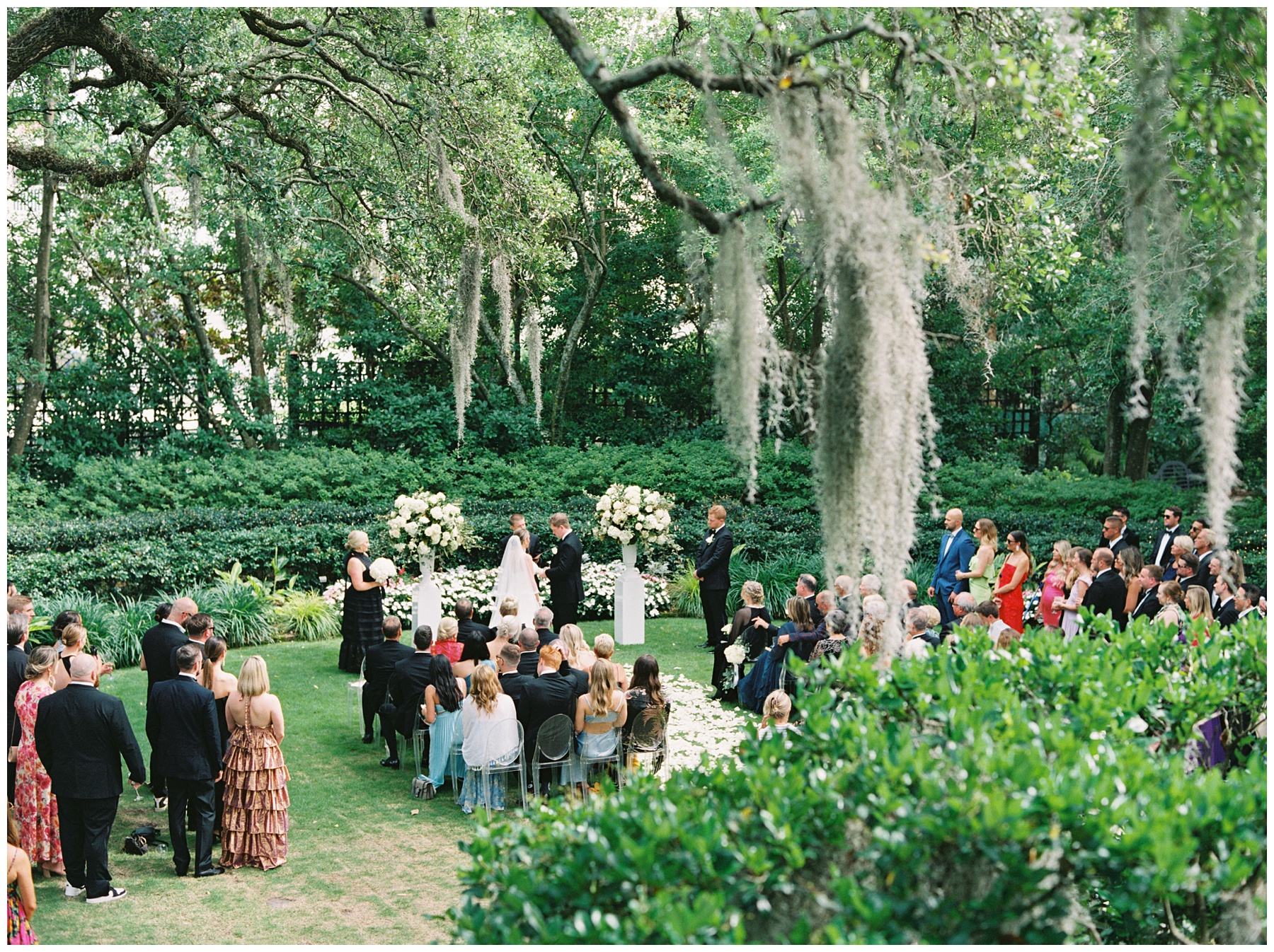 newlyweds change vows in garden under Spanish moss surrounded by white floral arrangements and white petals on lawn at the Governor Thomas Bennett House