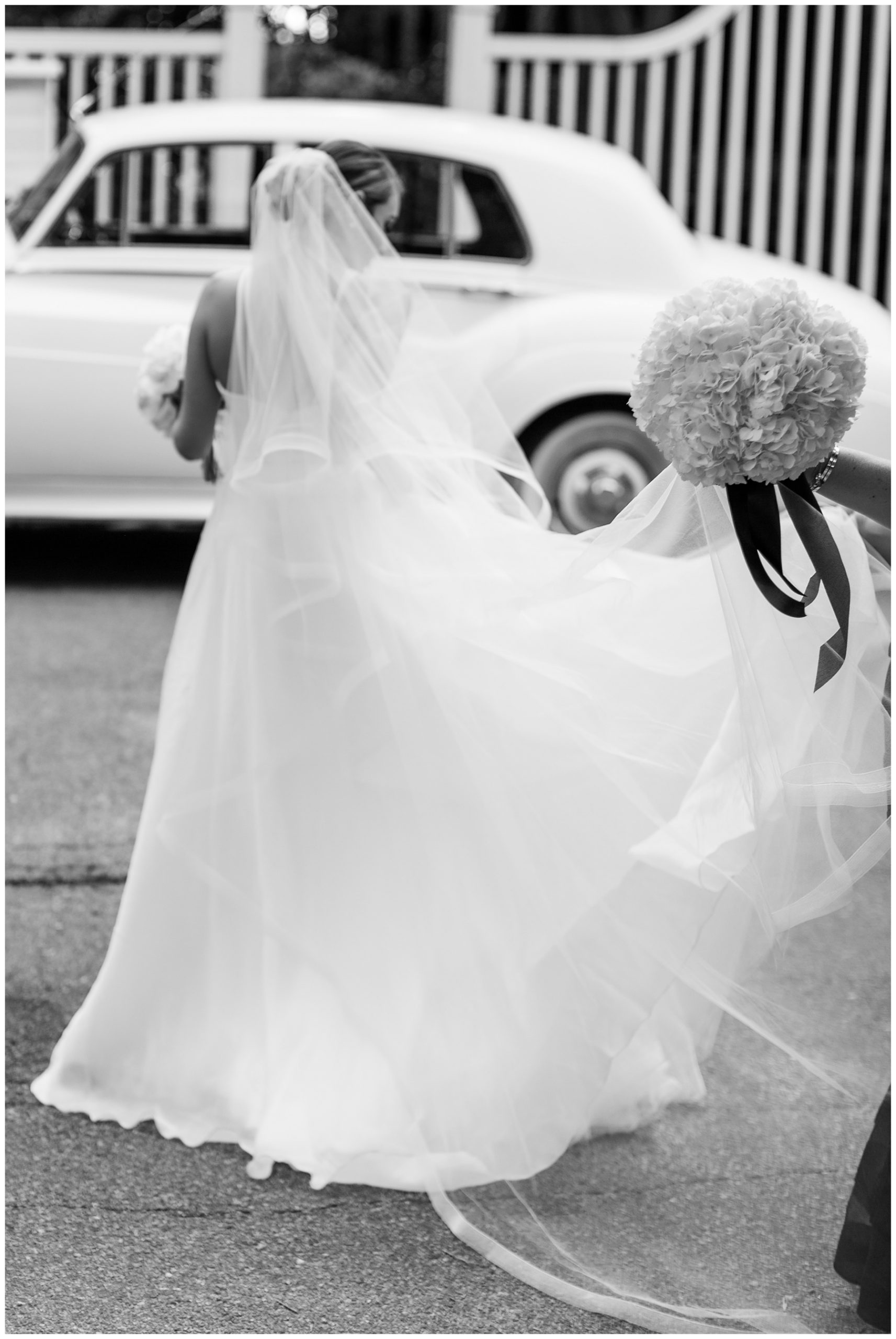 maid of honor holds up bouquet and back of bride's dress and veil walking towards classic white car in Charleston SC