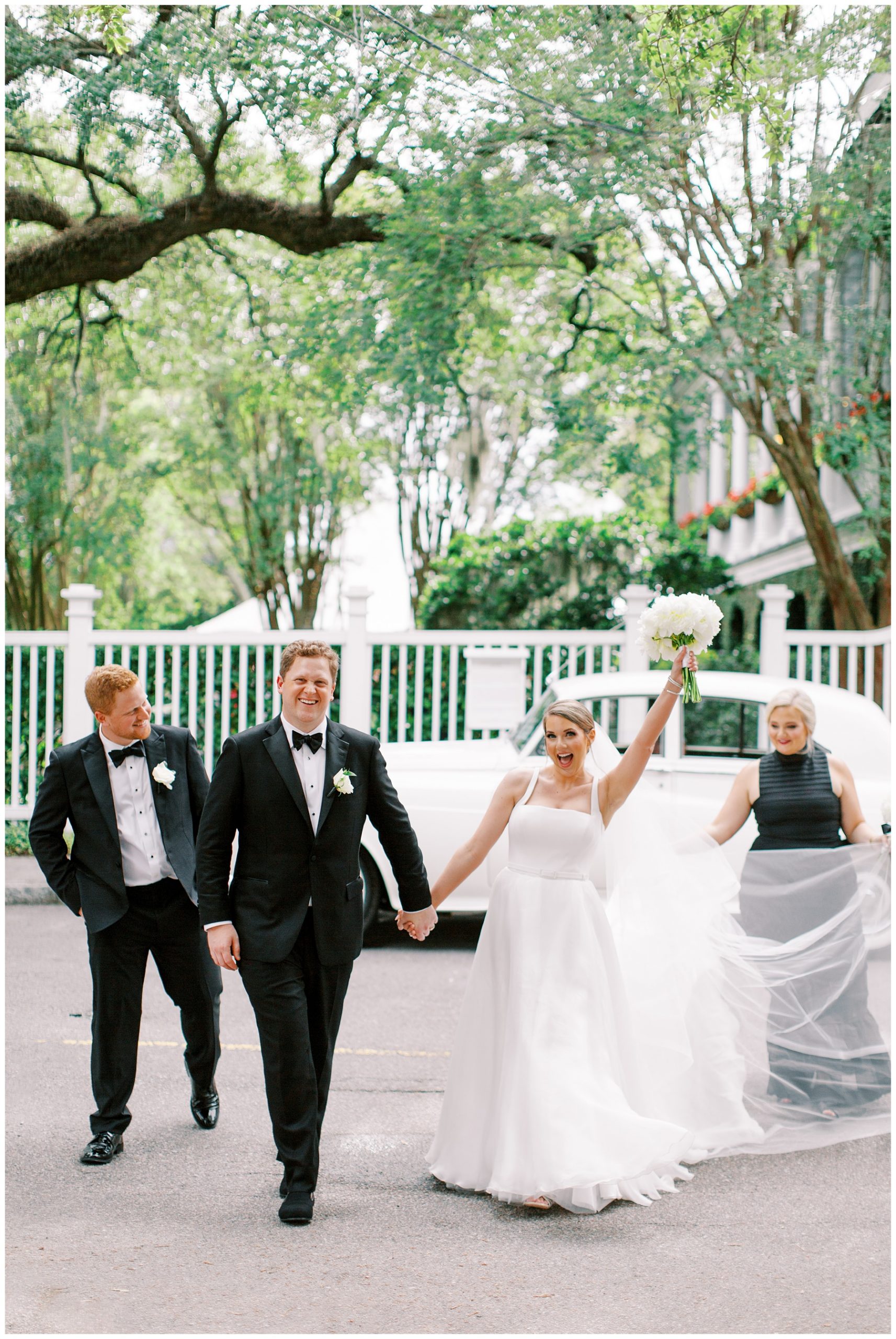 groom holds bride's hand walking across street in Charleston SC while she holds up bouquet of white flowers and maid of honor in black dress pulls out her veil