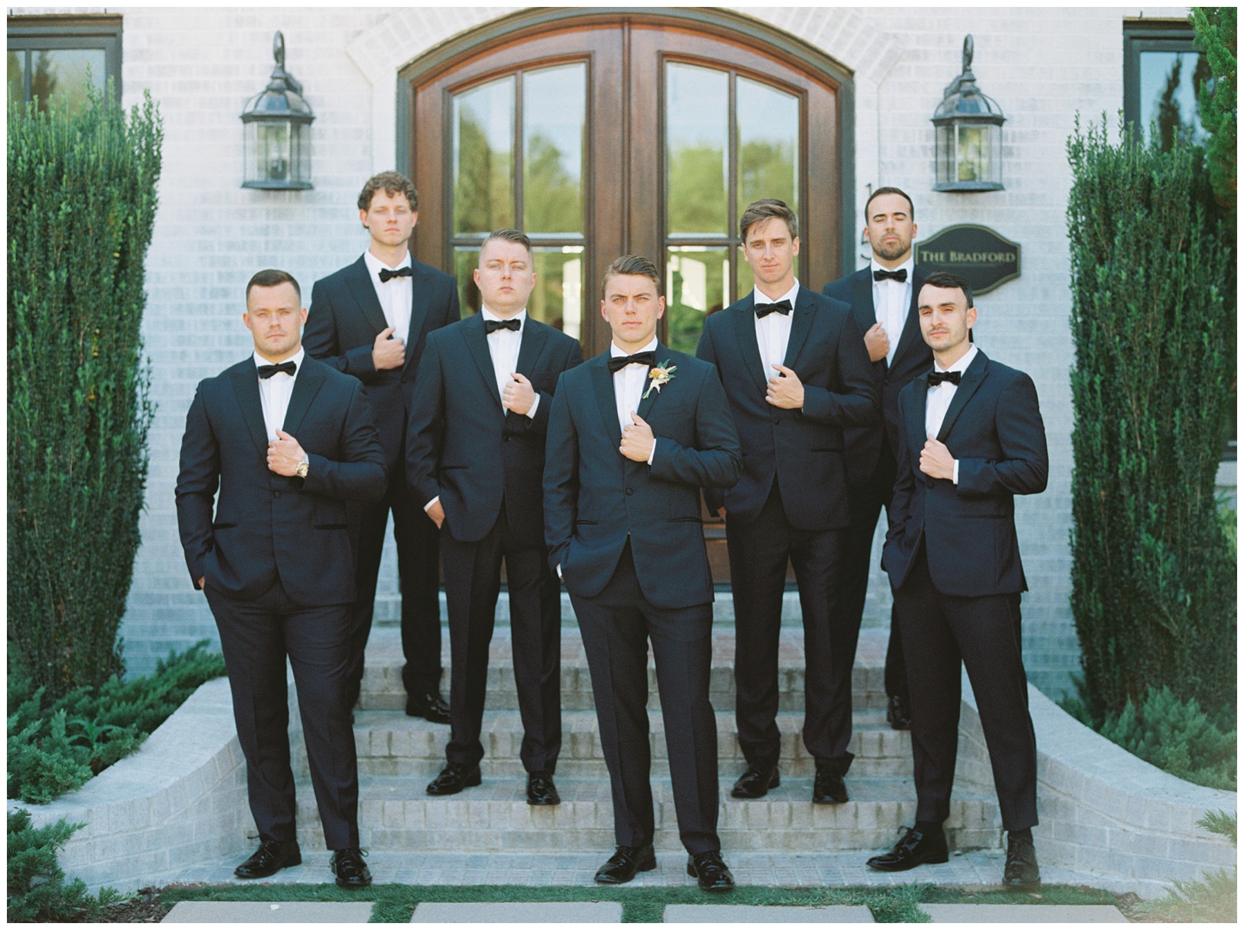 groom and groomsmen stand on brick steps outside The Bradford holding lapels of black and blue suits 