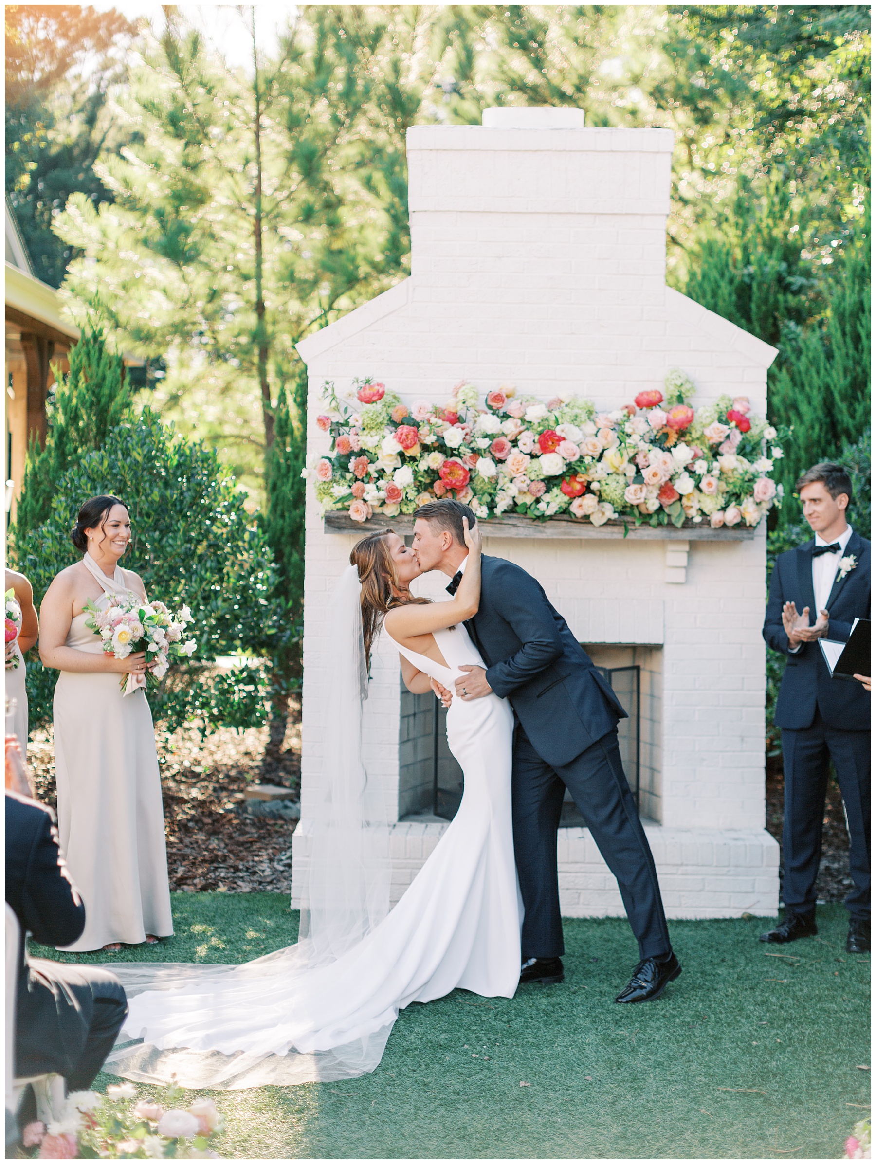 groom dips bride kissing her in front of white fireplace with pink and white flowers on mantel during ceremony 