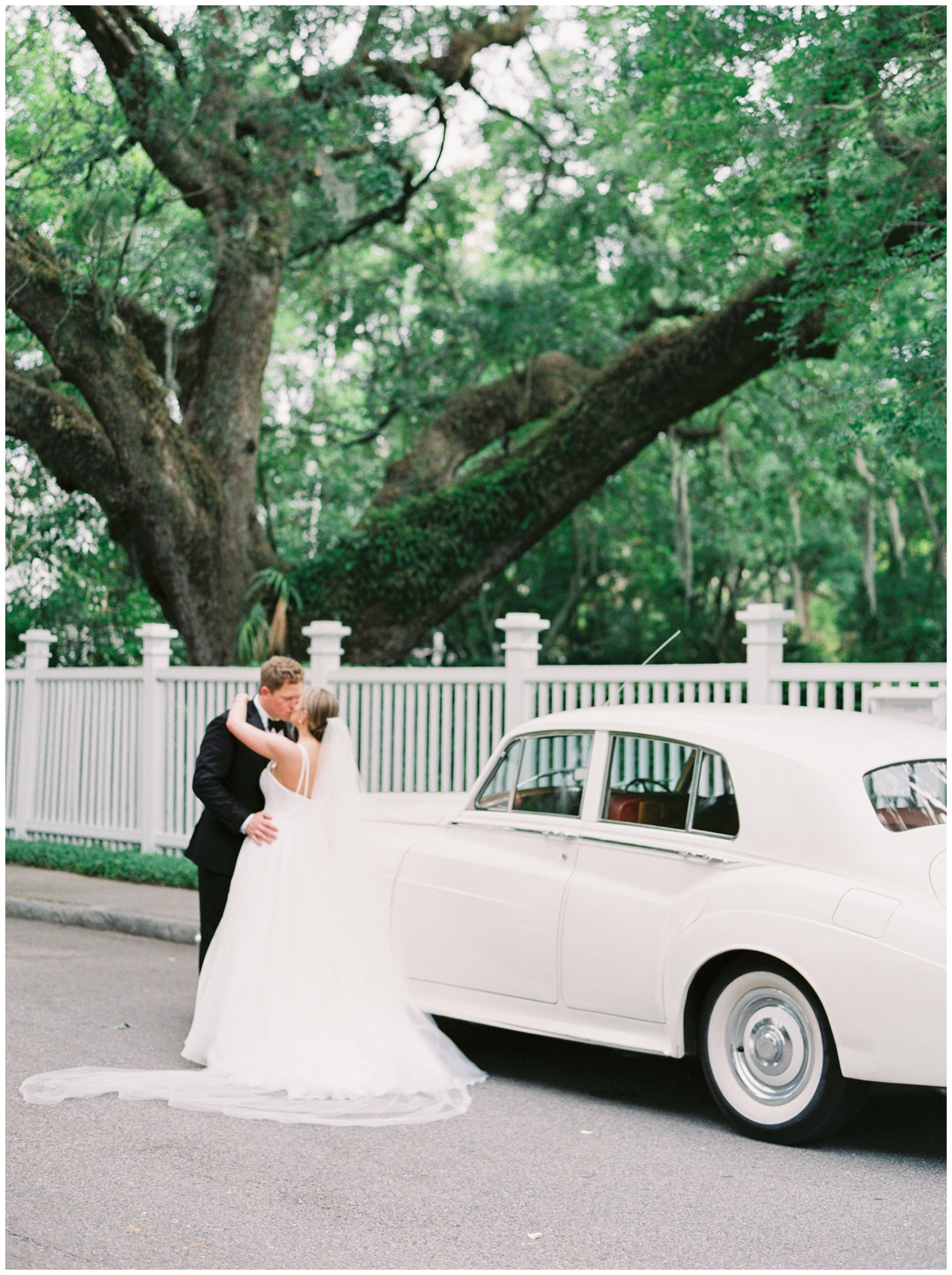 newlyweds kiss by white car beside white picket fence in Charleston SC