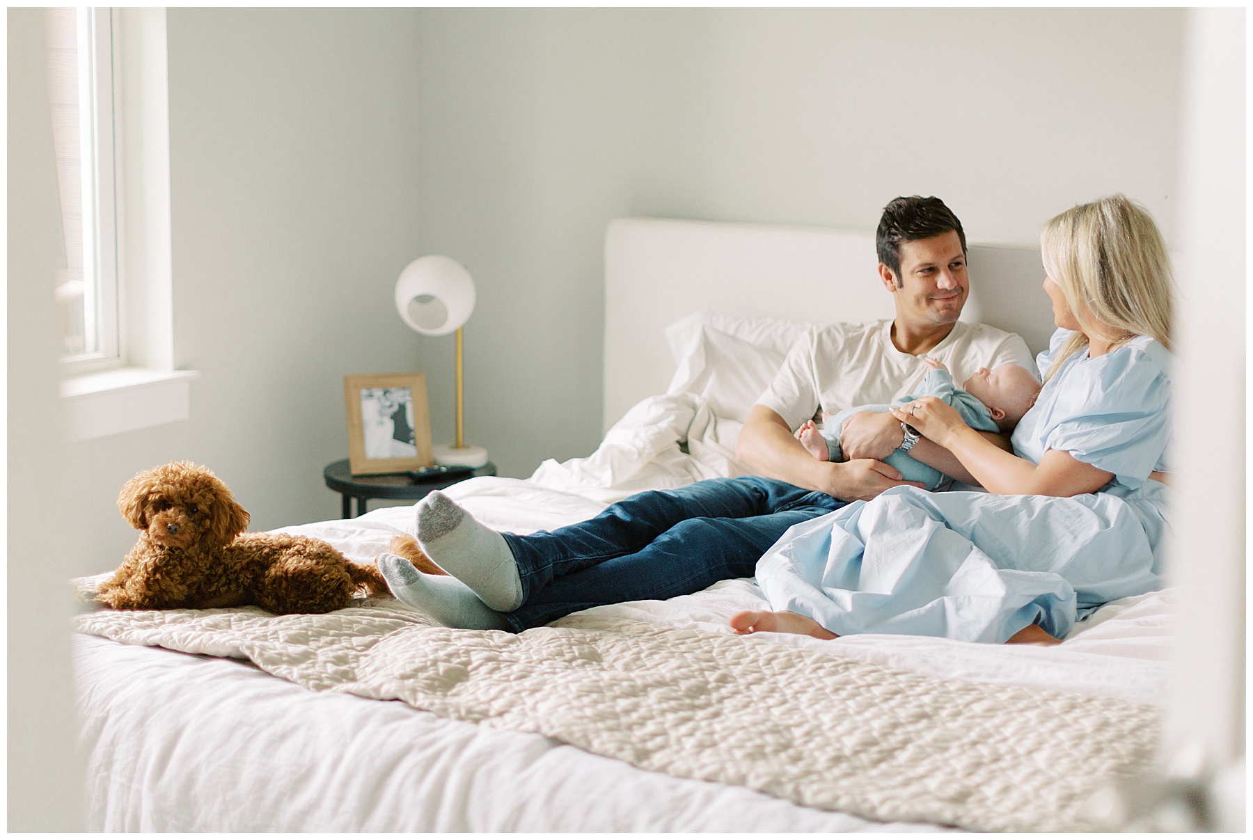 parents snuggle with son with dog on the foot of their bed during in-home newborn session