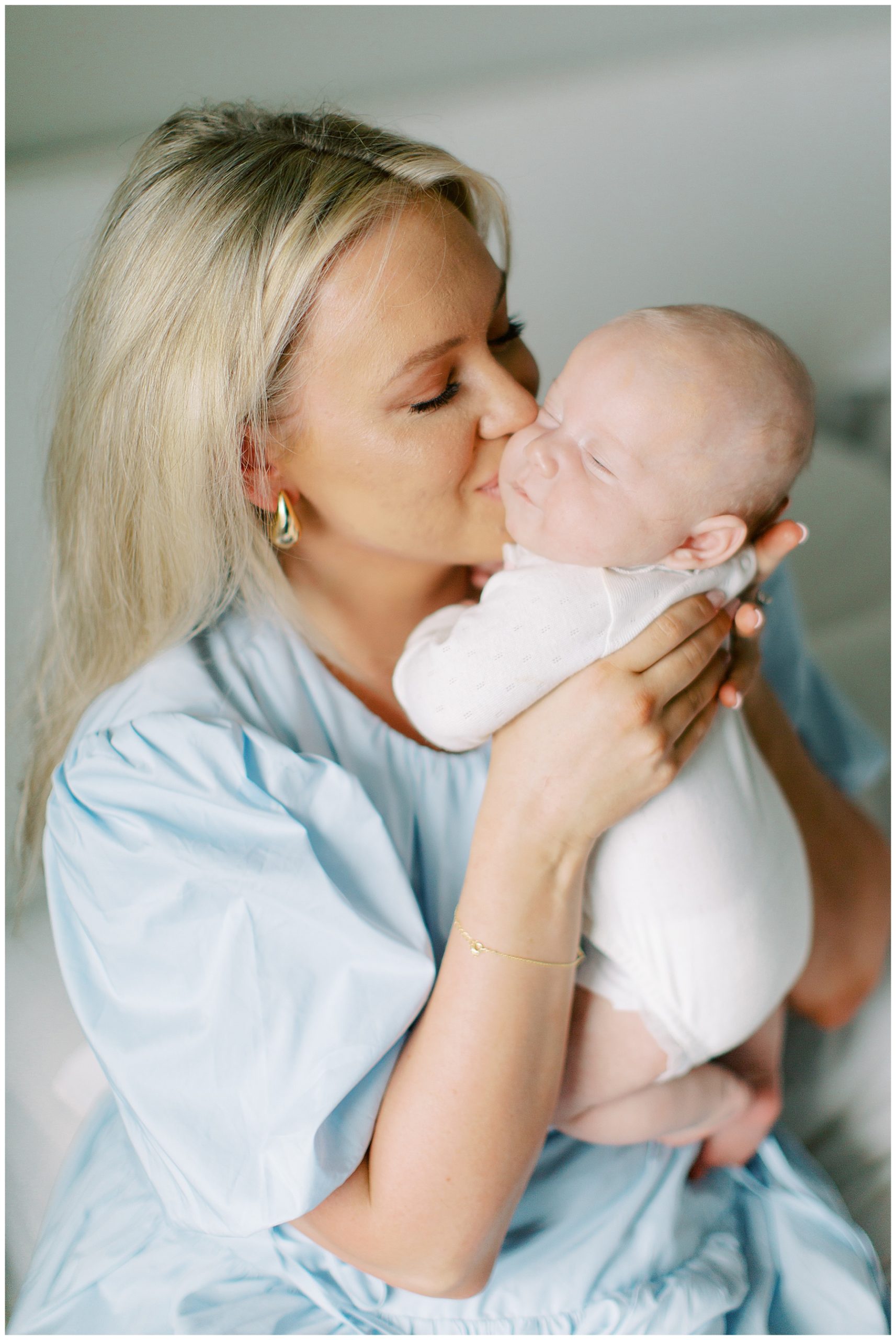 blonde woman in blue dress kisses baby's cheek in white knit outfit during Charlotte in-home newborn session