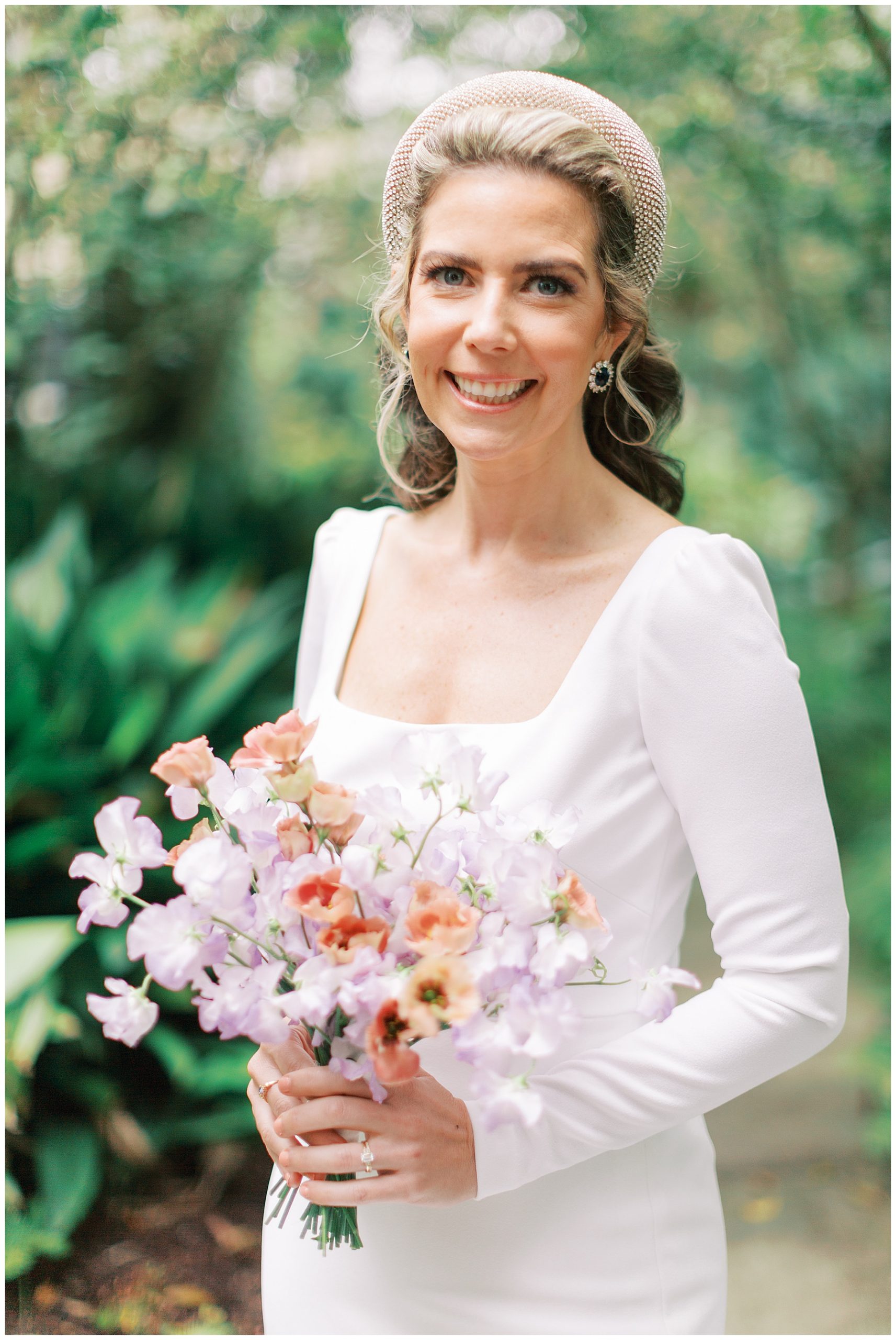 blonde bride in long sleeve white gown holds bouquet of purple and coral wildflowers 