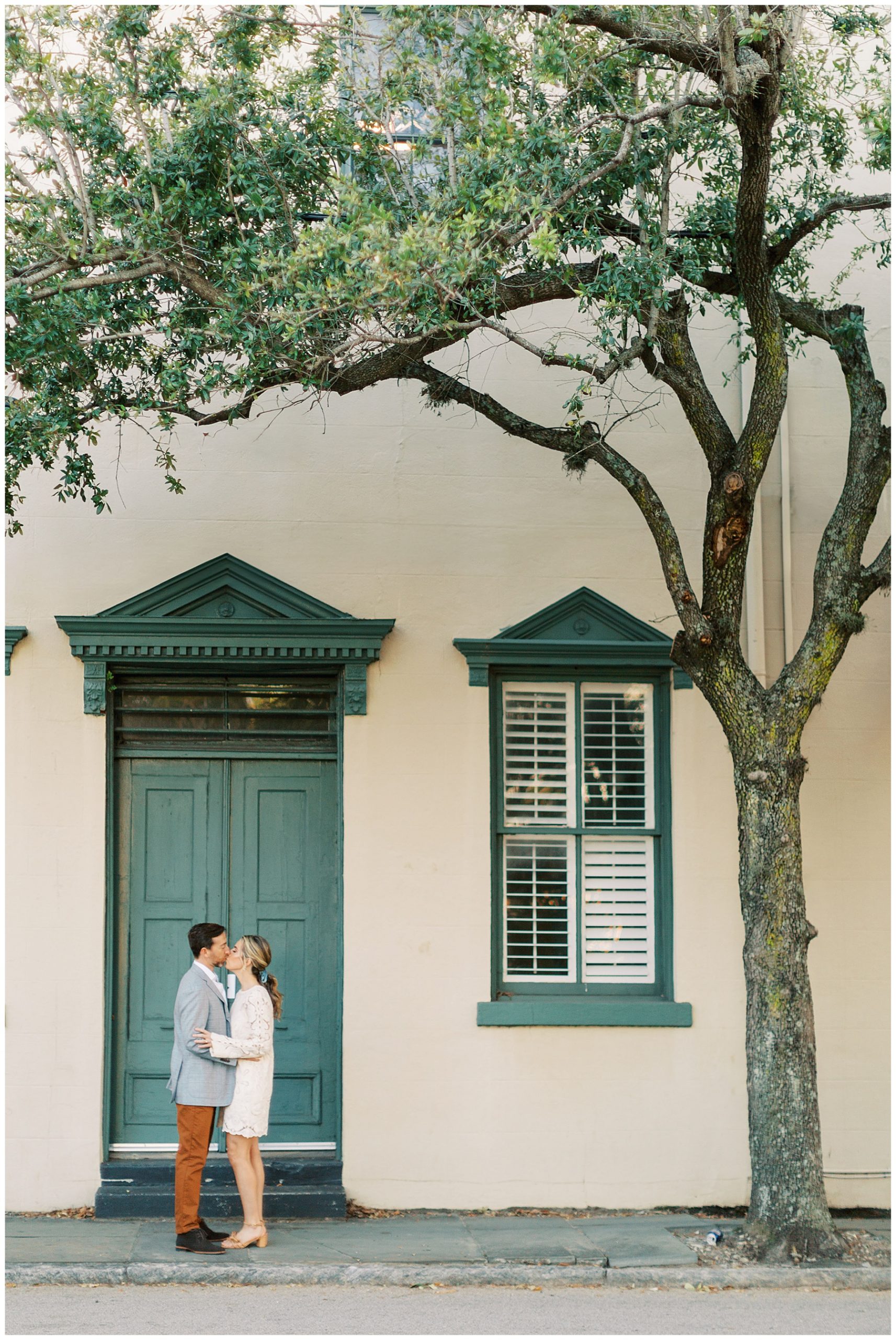 woman in white lace gown hugs man in blue suit jacket in front green door of Charleston home 
