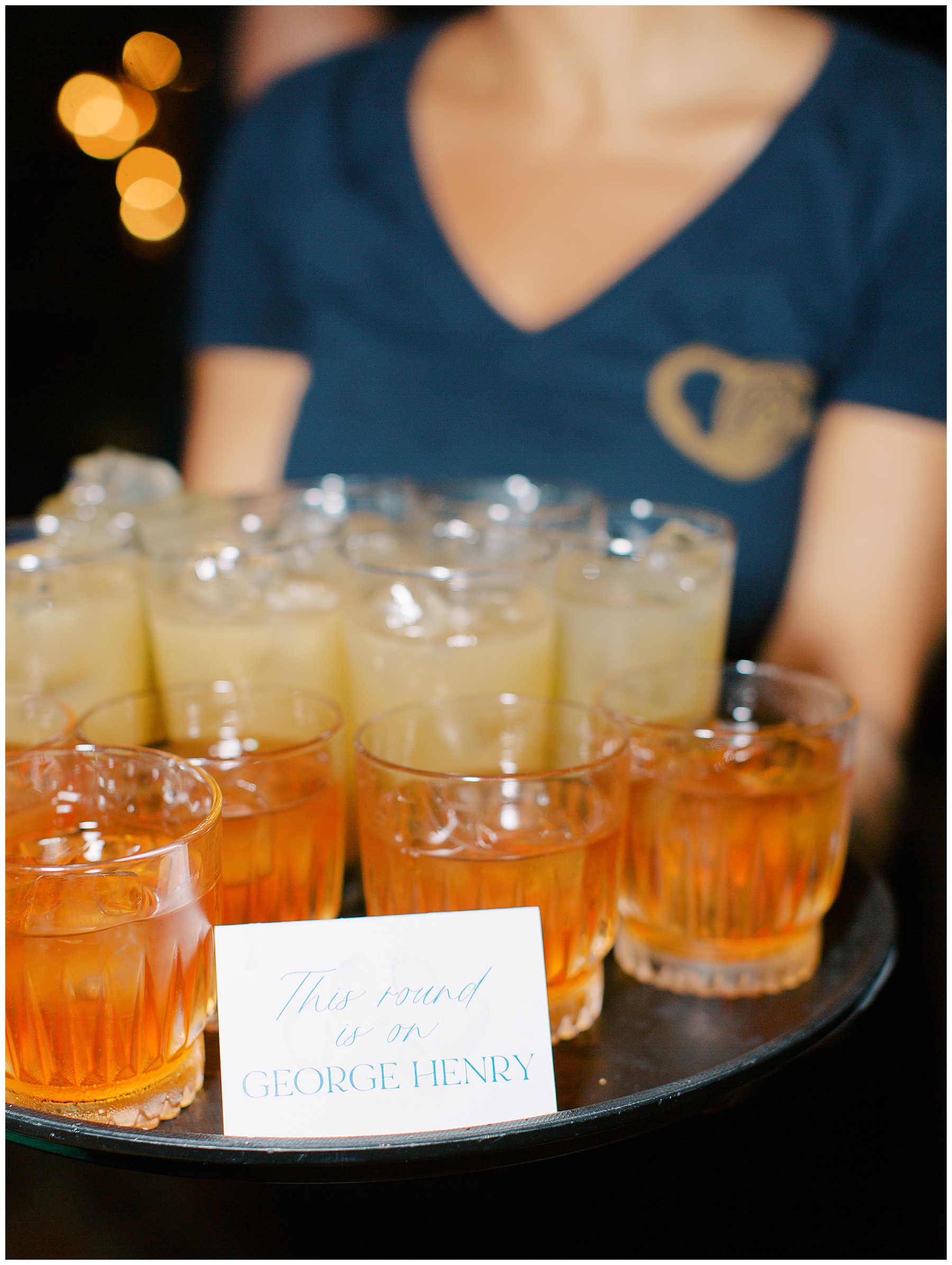 woman holds tray of lemonade and bourbon with card that says "this round is on George Henry" 