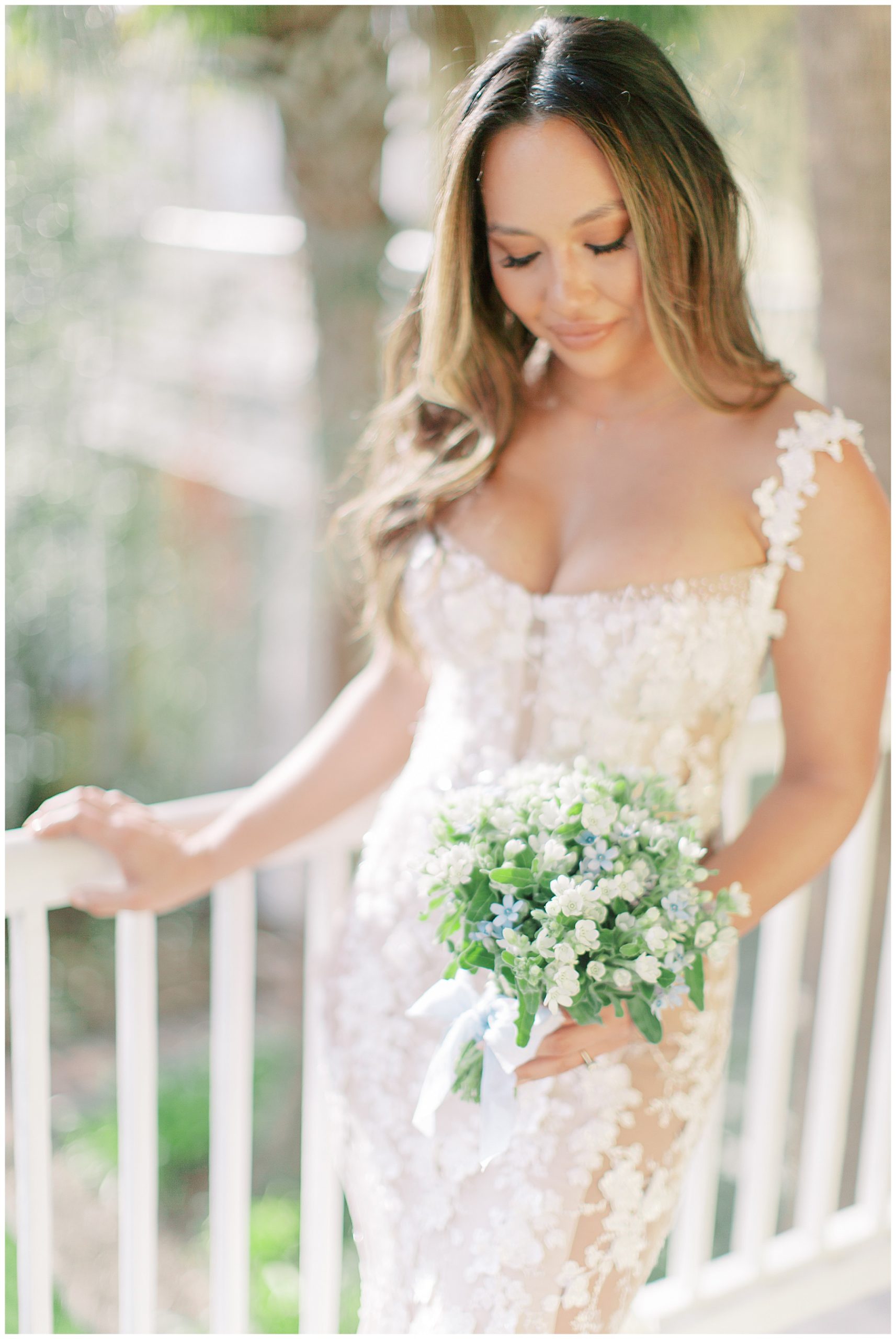 bride looks down at small bouquet of white and blue flowers with white ribbon while holding railing of porch outside the William Aiken House