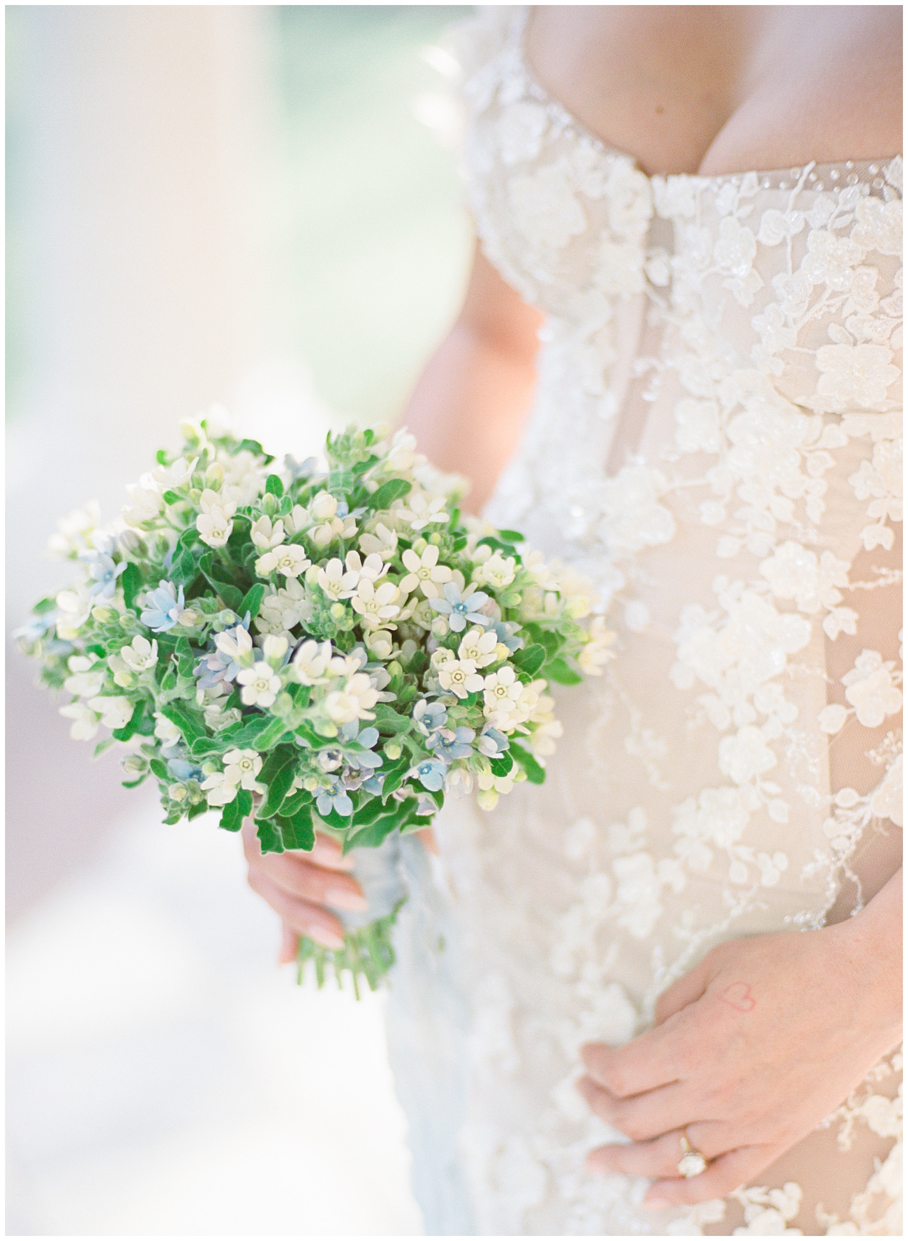 bride holds small bouquet with white and blue flowers in front of lace topped wedding gown in Charleston SC