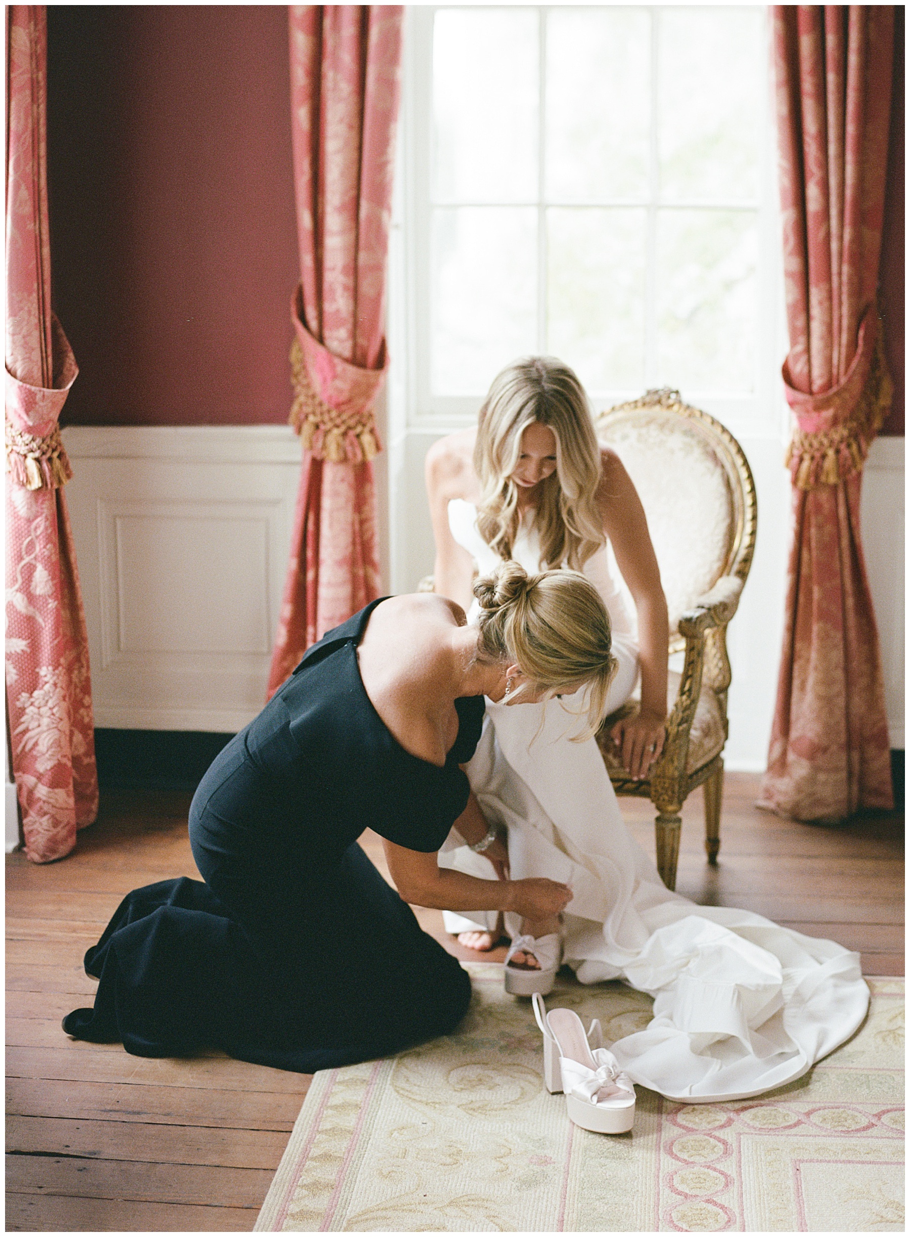 woman kneels to help bride with white shoes on wedding morning 