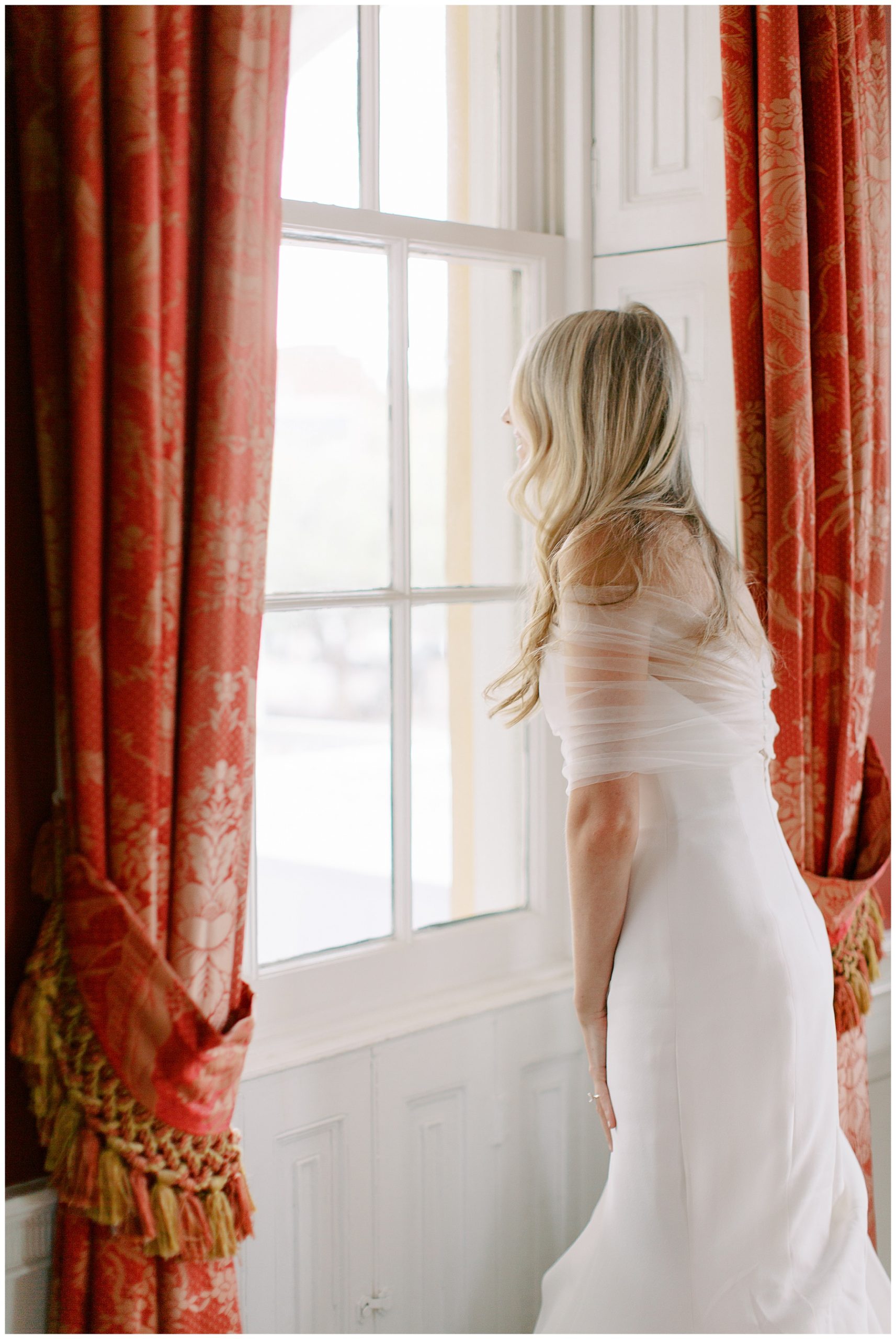 blonde bride with tulle wrap around her looks out window of the William Aiken House
