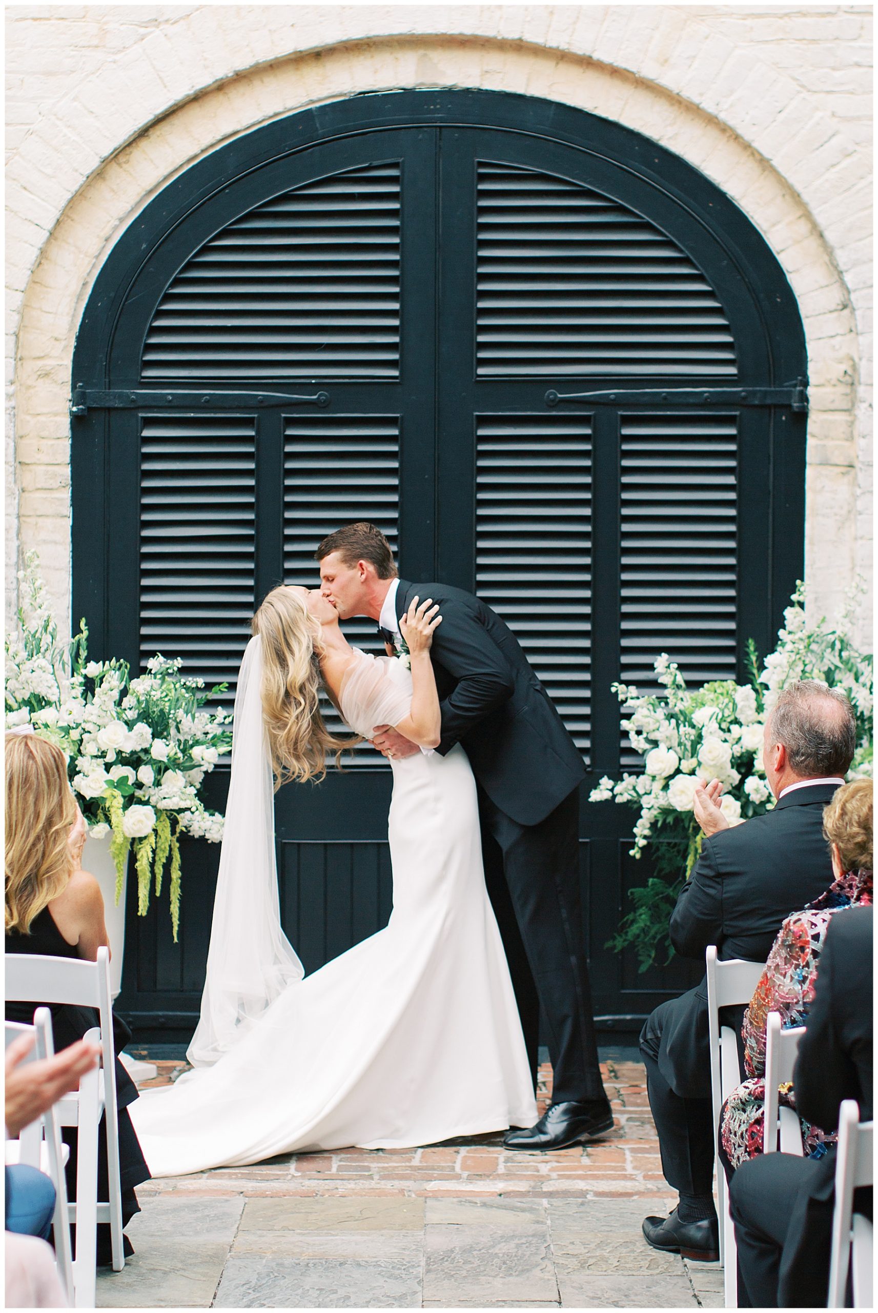 groom dips bride kissing her in front of black shuttered doors at the William Aiken House during ceremony 