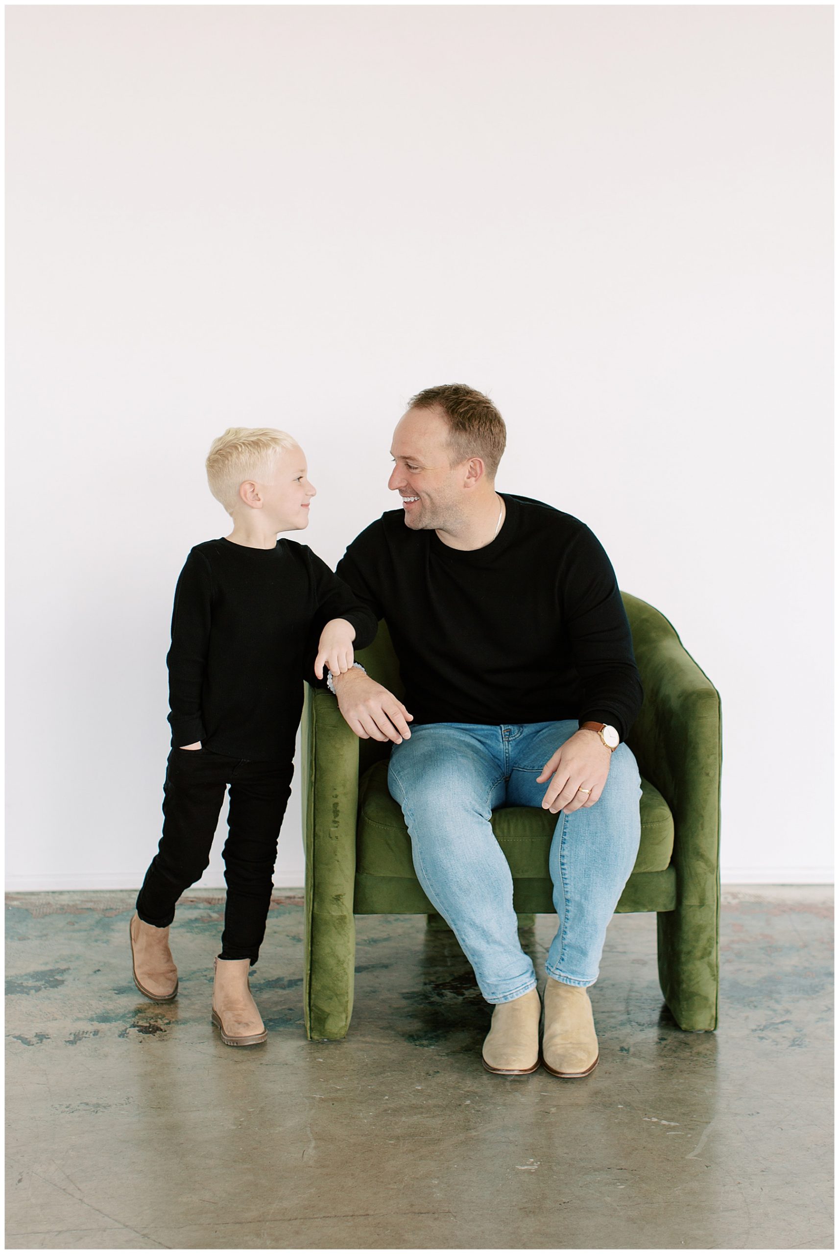 father in black shirt and jeans sits in green chair talks to son standing next to him during Charlotte studio family session