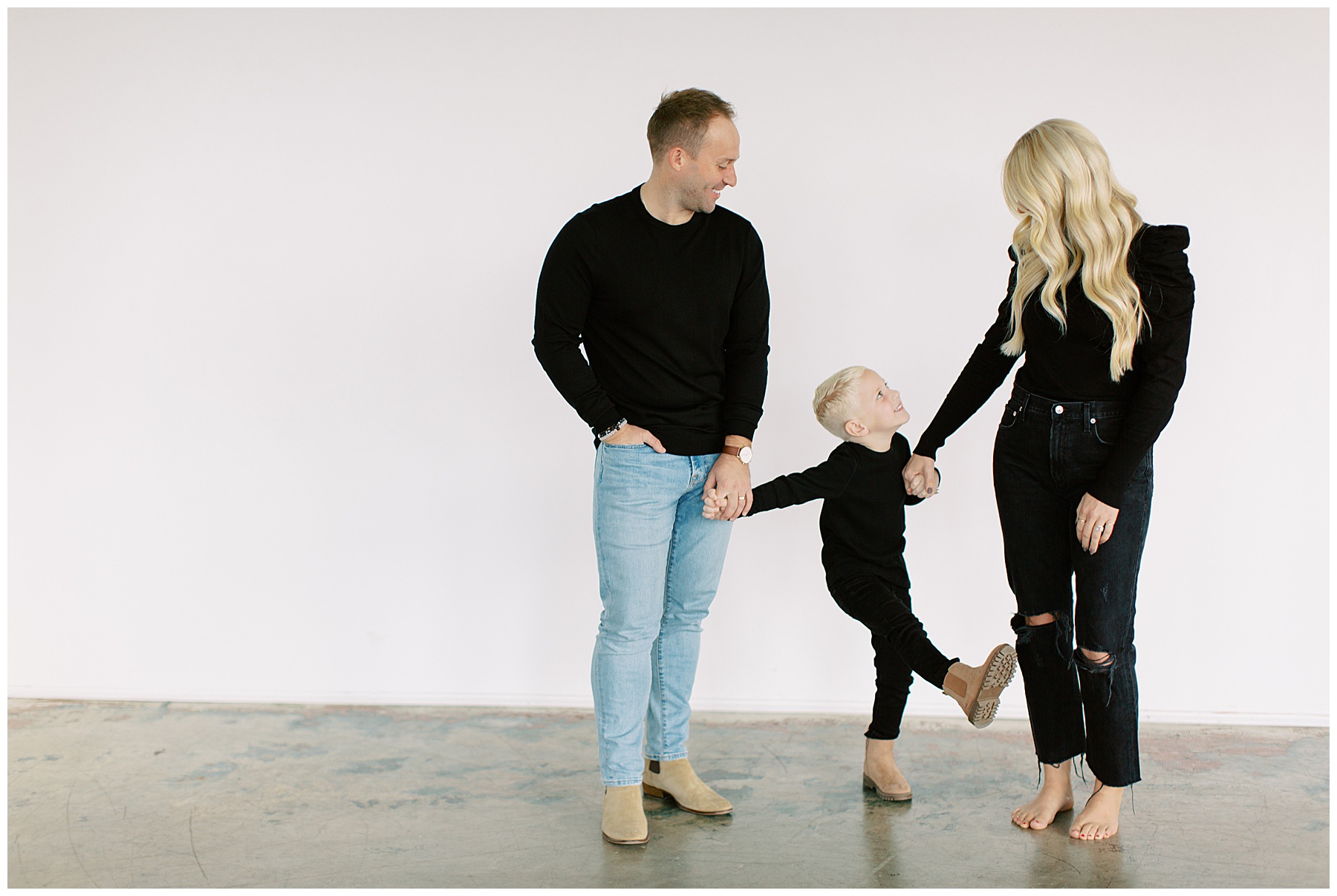 married couple stands holding their son's hands during Charlotte studio family session in front of white wall