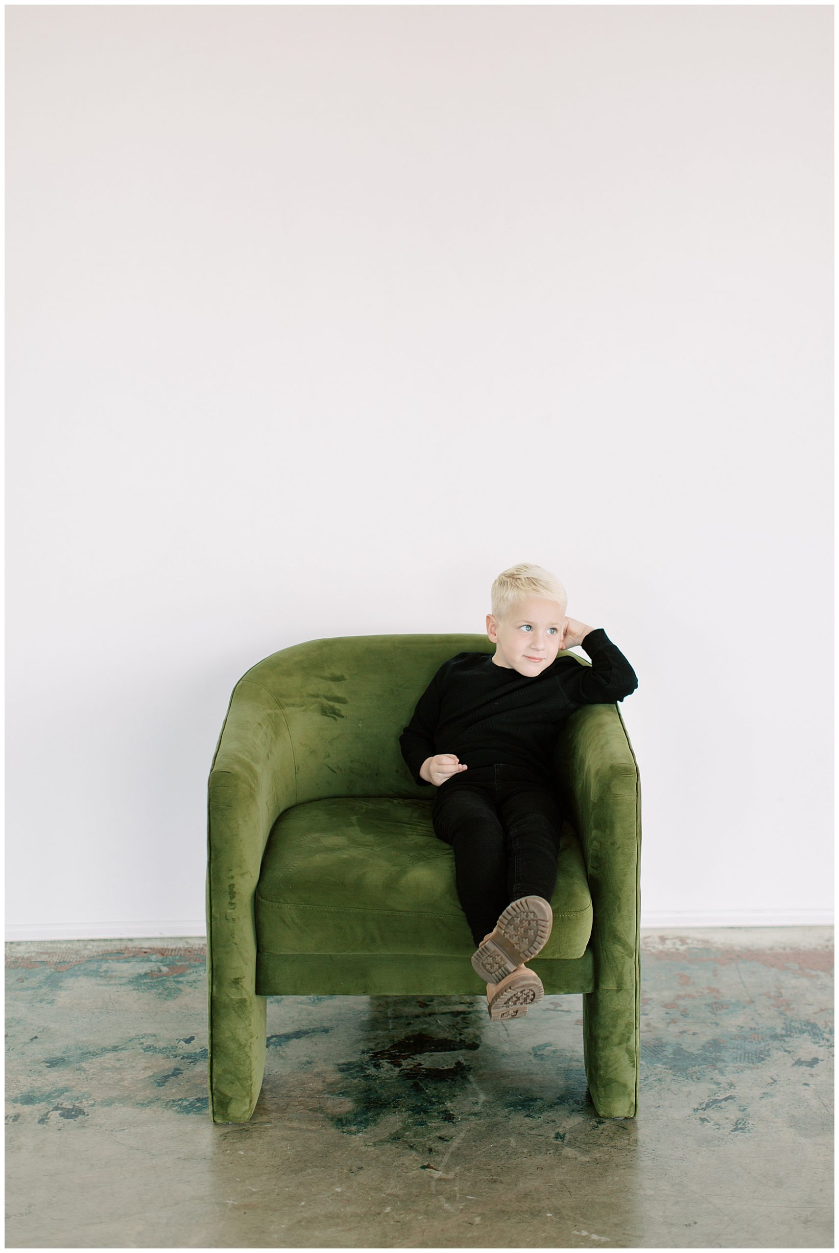 boy with blonde hair and blue eyes sits in green chair wearing black outfits in tan boots