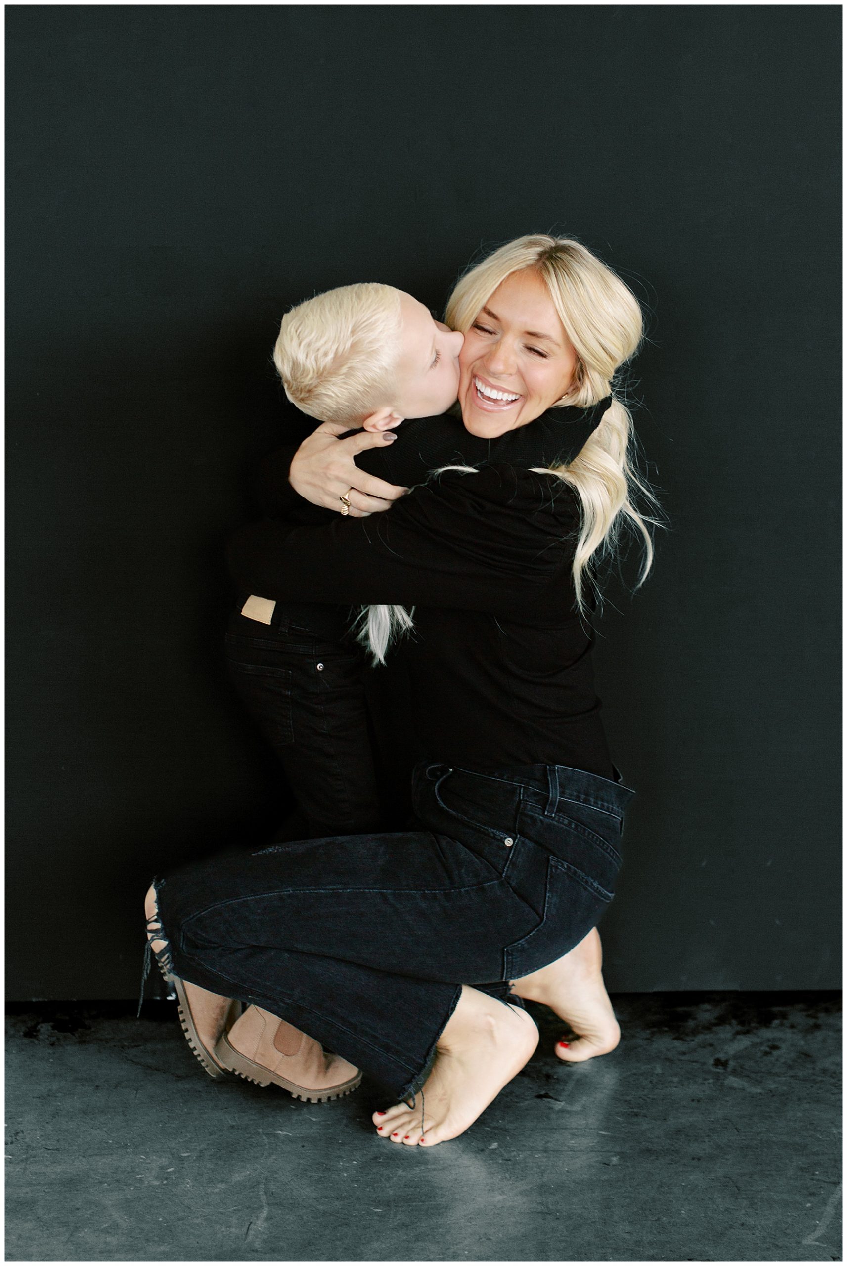 mom kneels while son hugs her making her laugh during studio family photos 