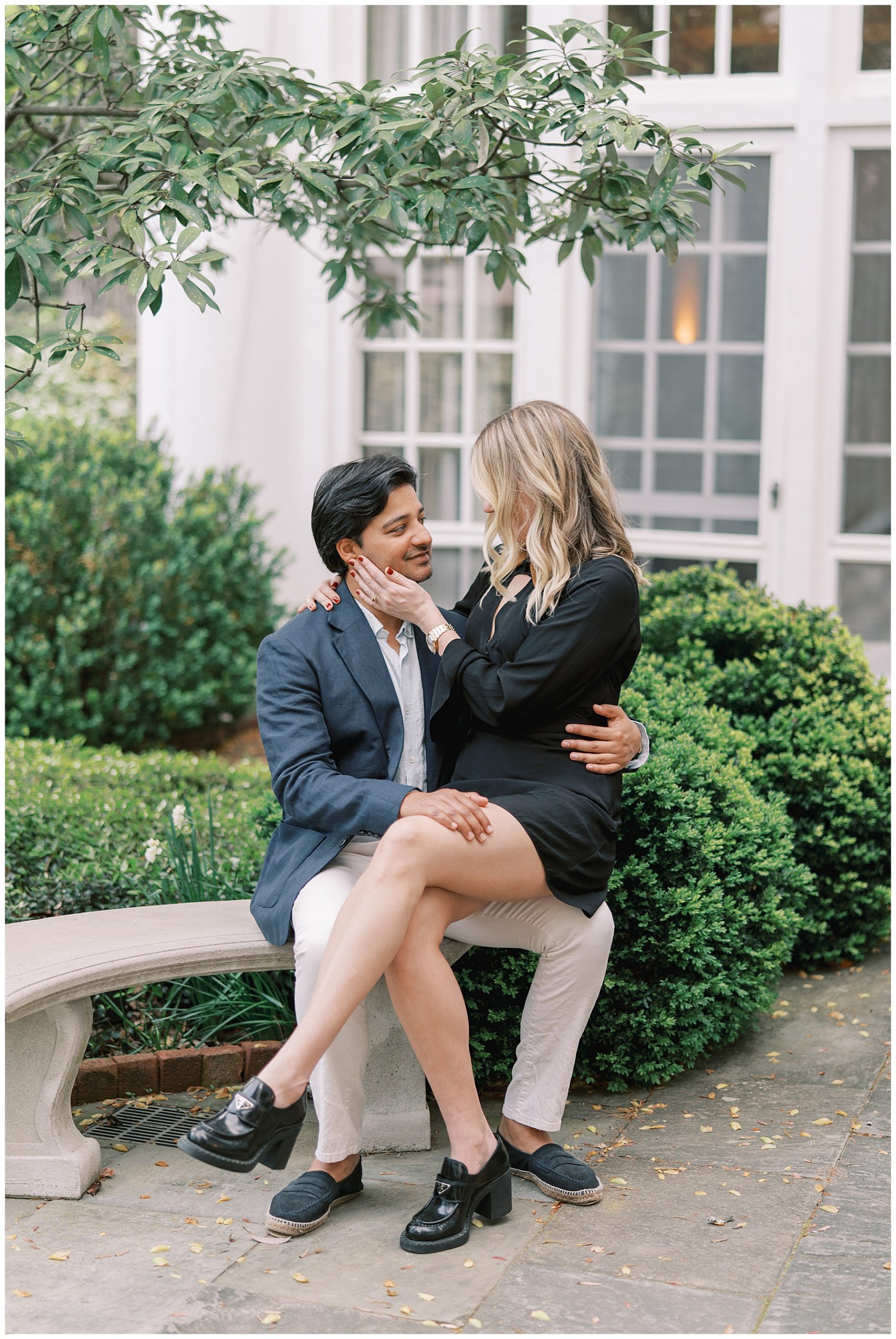 woman in black dress sits on man's lap during engagement portraits in the gardens of Duke Mansion