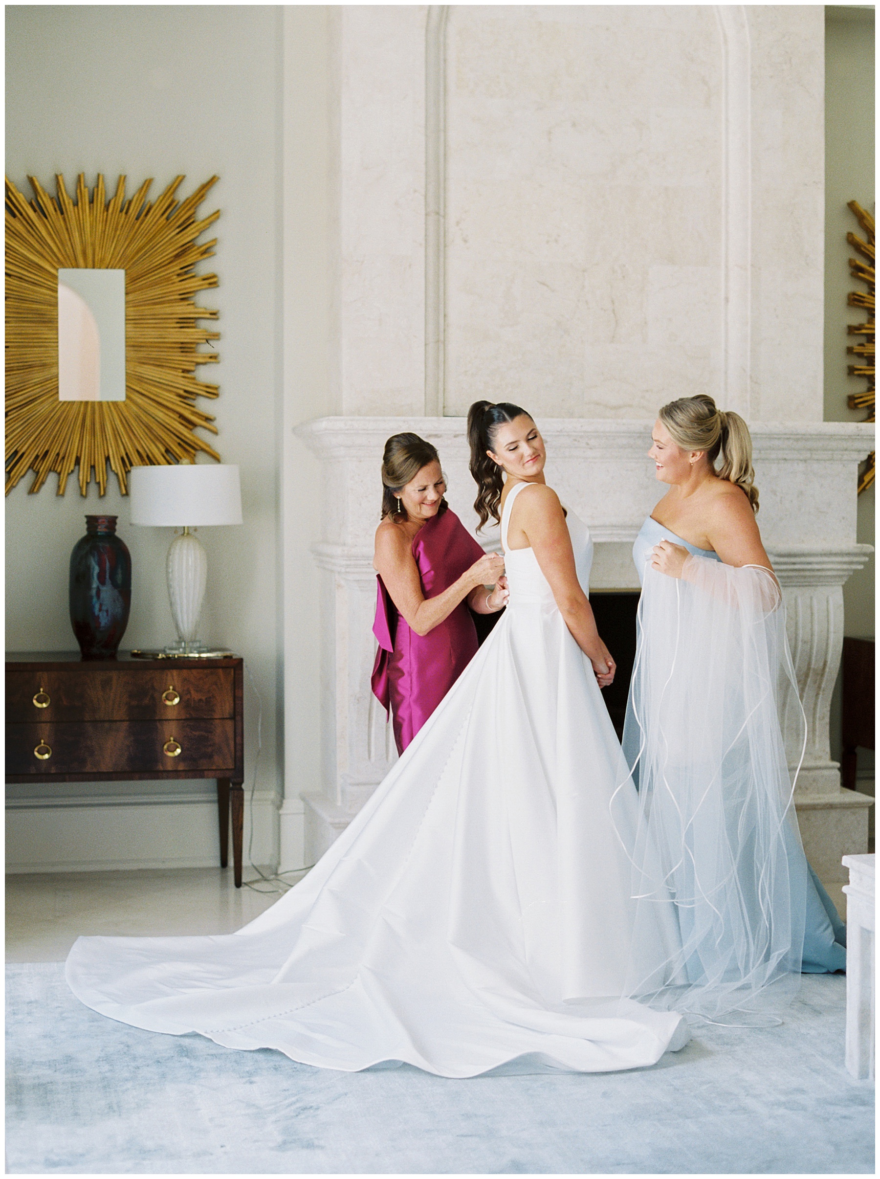 mother buttons up bride's dress while bridesmaid holds veil in suite at Grey Oaks Country Club