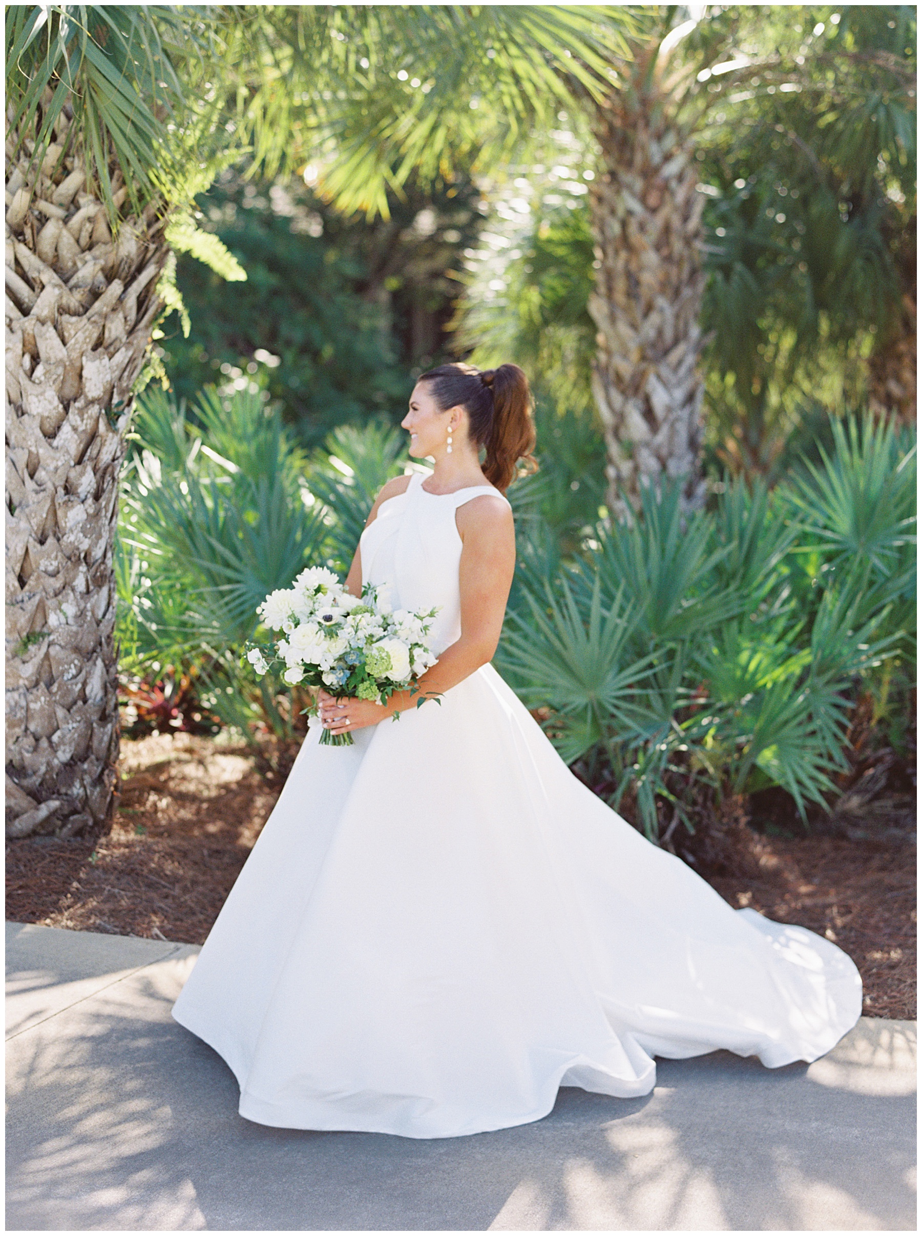 bride stands in chic wedding gown holding bouquet of white flowers by palm trees at Grey Oaks Country Club