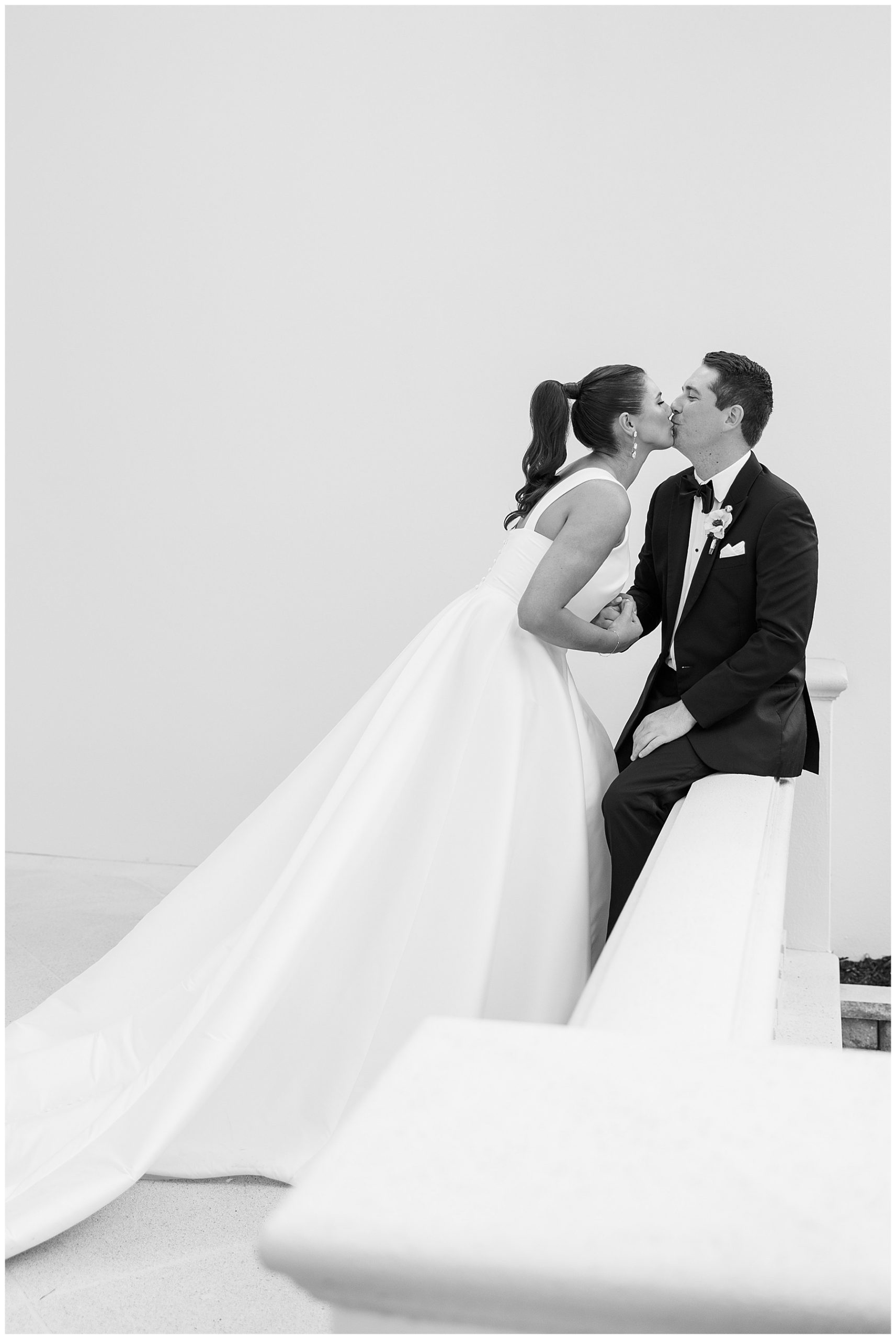 bride leans to kiss groom sitting on white railing at Grey Oaks Country Club