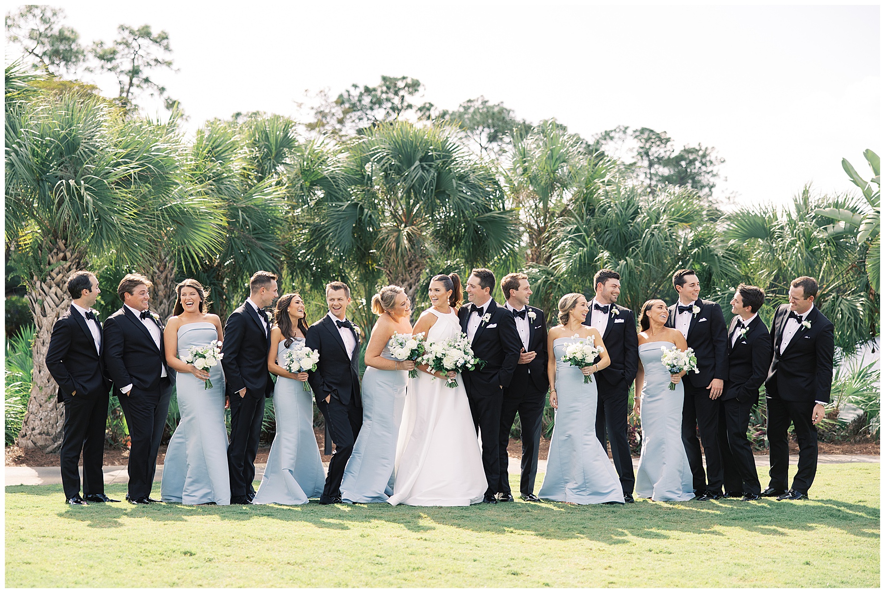 bride smiles at bridesmaids standing with groomsmen in black suits in front of palm trees at Grey Oaks Country Club