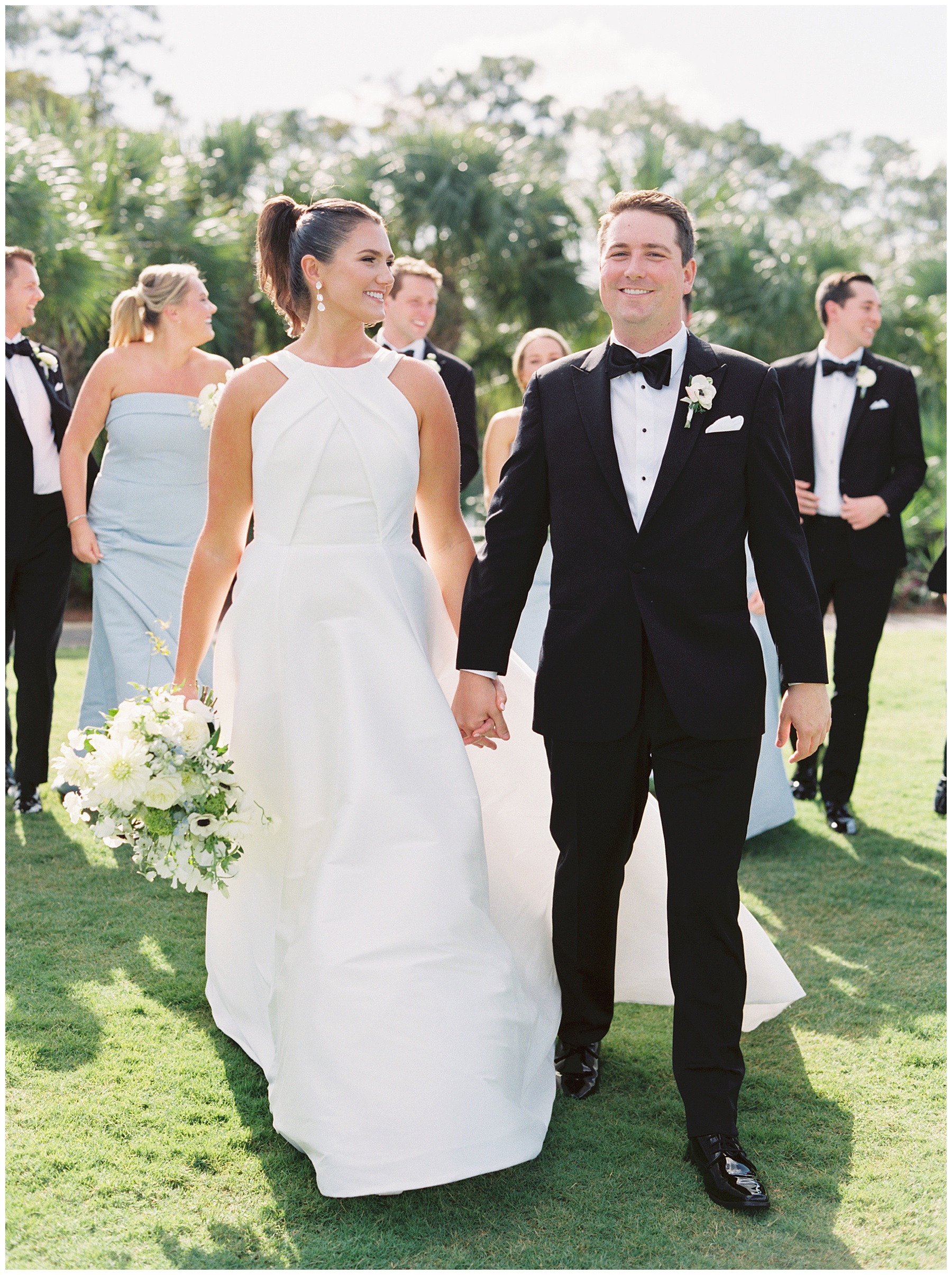 newlyweds hold hands walking in front of wedding party in blue gowns and tuxes 