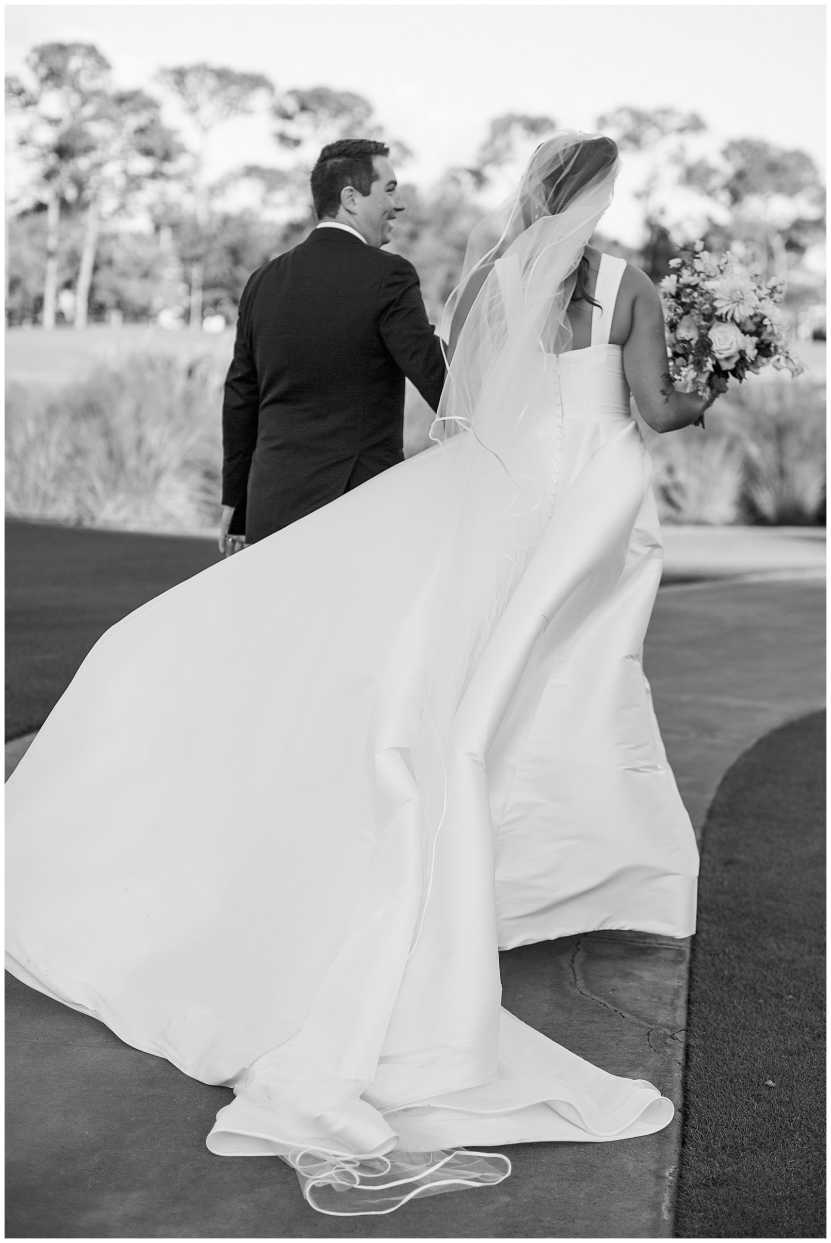 bride and groom hold hands walking down pathway with her veil blowing in the wind