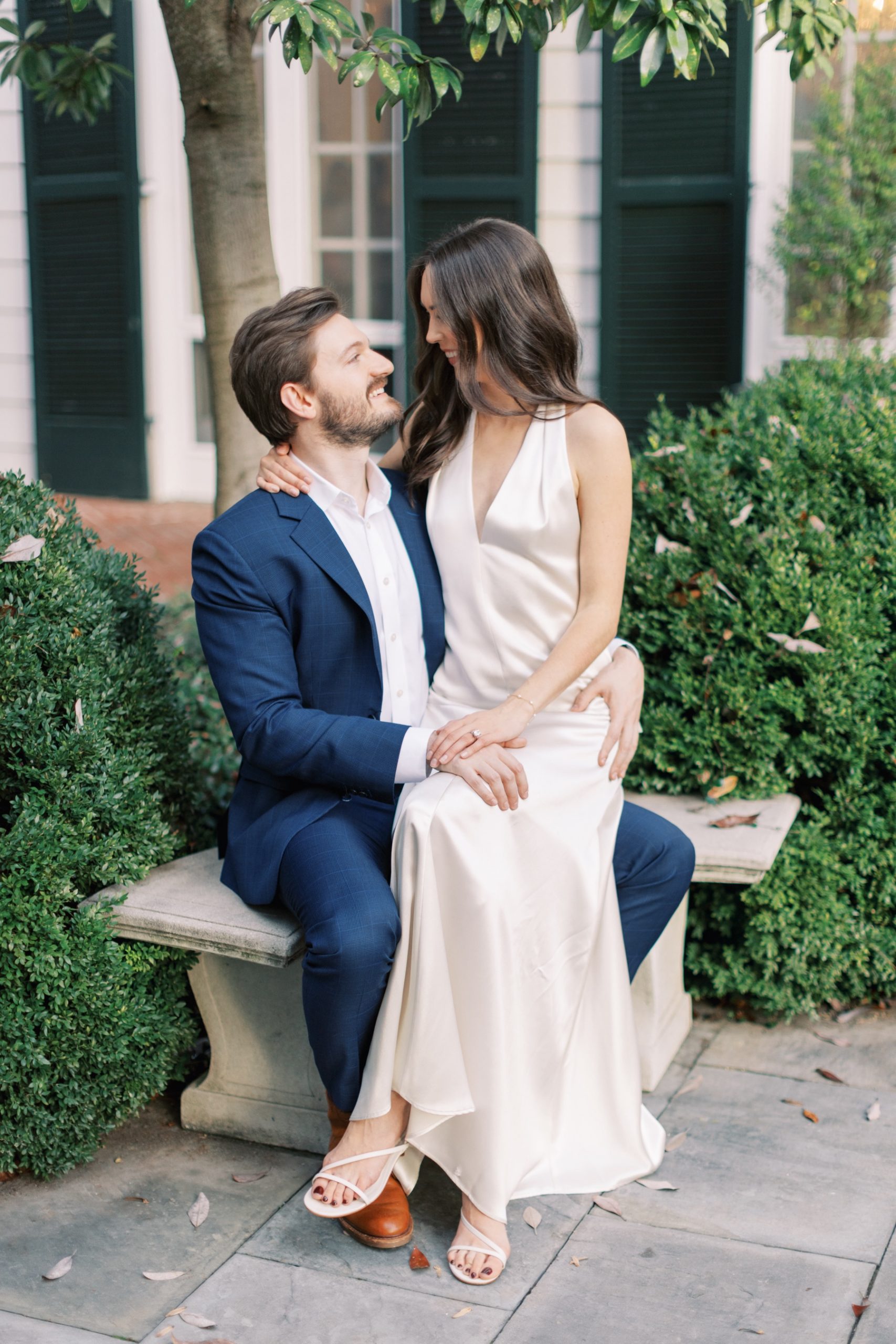 woman in satin white dress sits on man's lap on stone bench during engagement session at Duke Mansion