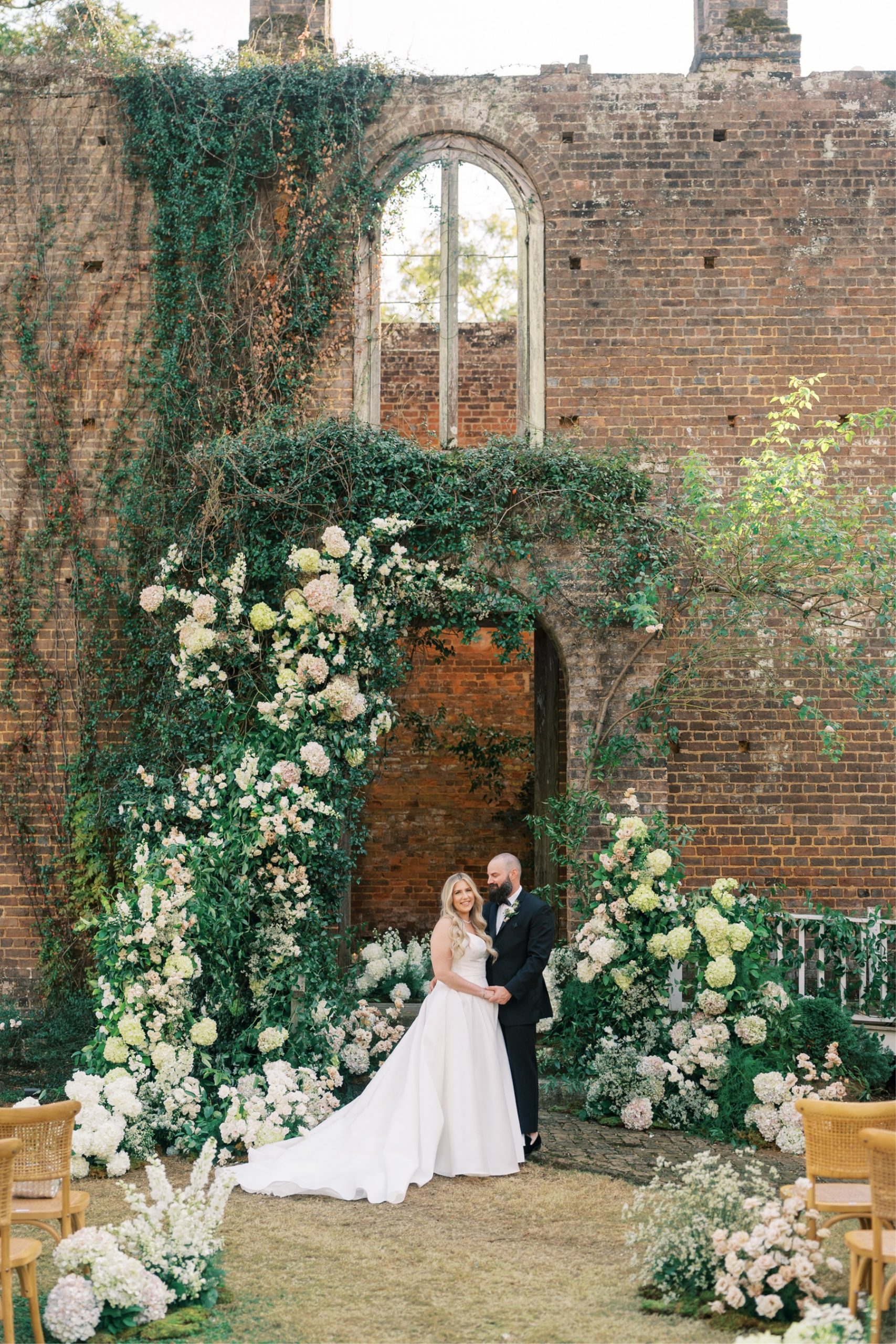 Bride and groom standing under the archway covered in flowers in front of the ruins of a building at the Barnsley Resort