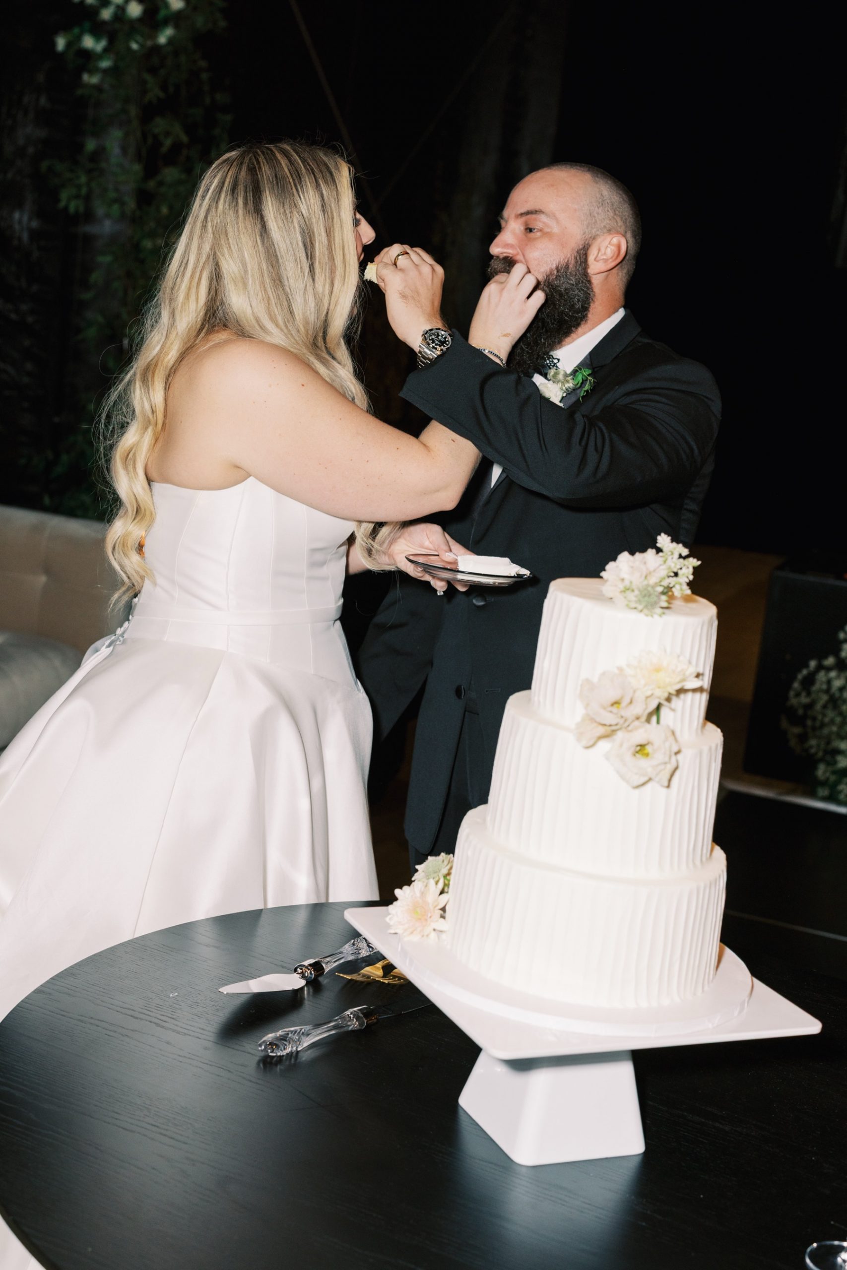 Bride and groom feeding each other cake with simple 3 tiered white wedding cake in the foreground