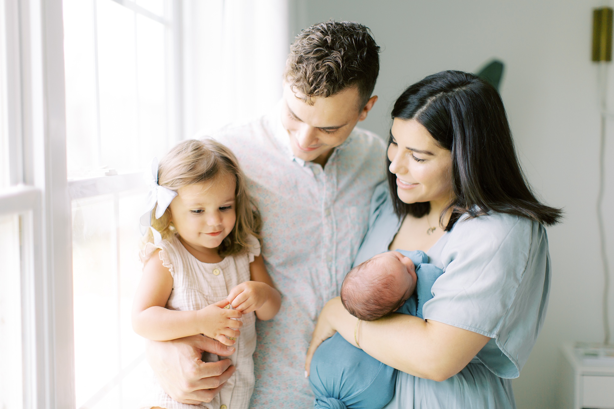 family of four poses by window during Charlotte lifestyle newborn session