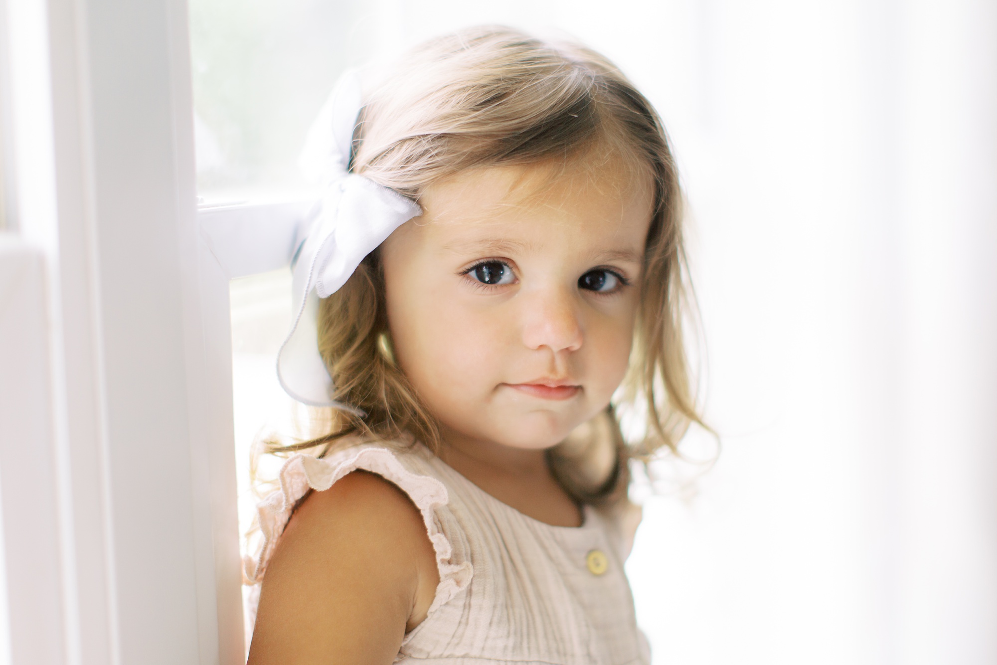 little girl with white bow poses by window during lifestyle photos at home