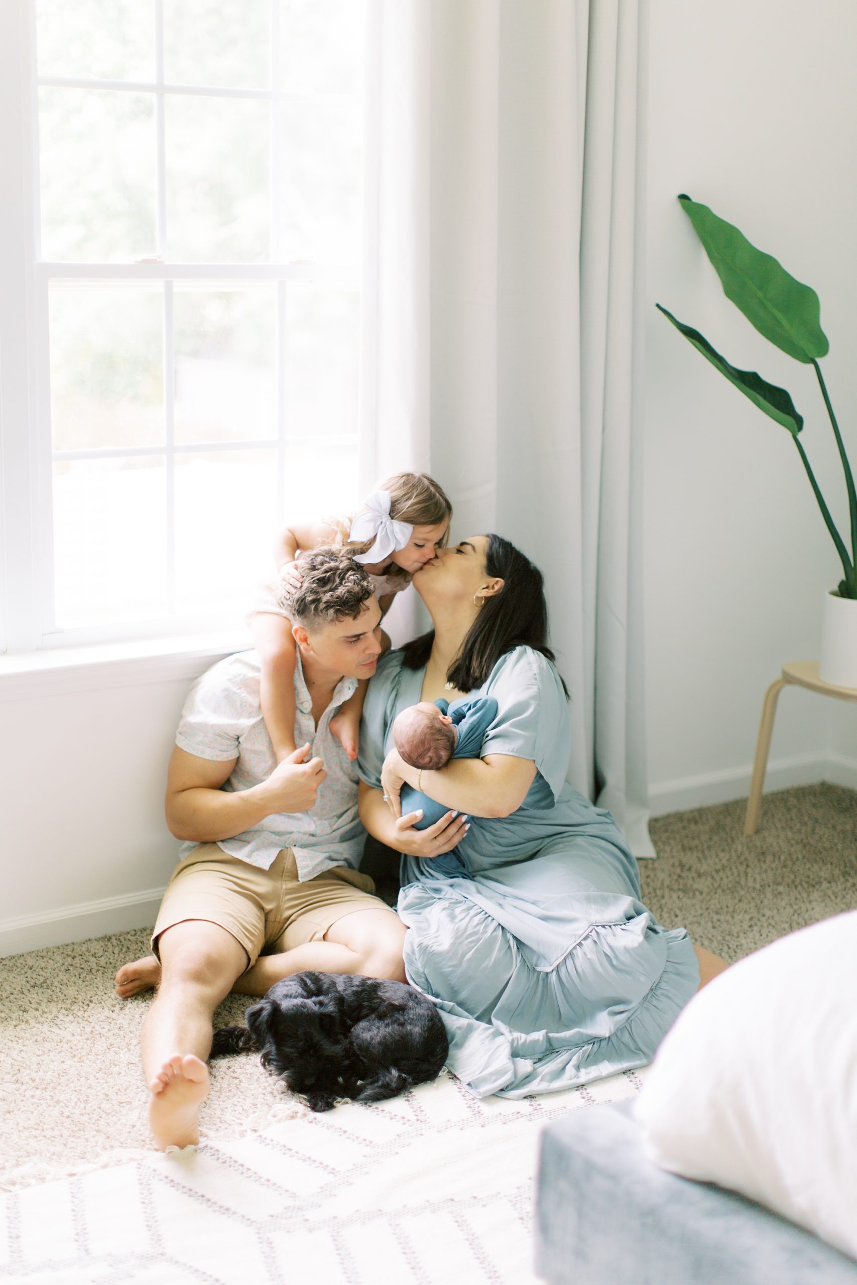 big sister sits on dad's shoulders and leans to kiss mom during Charlotte lifestyle newborn session