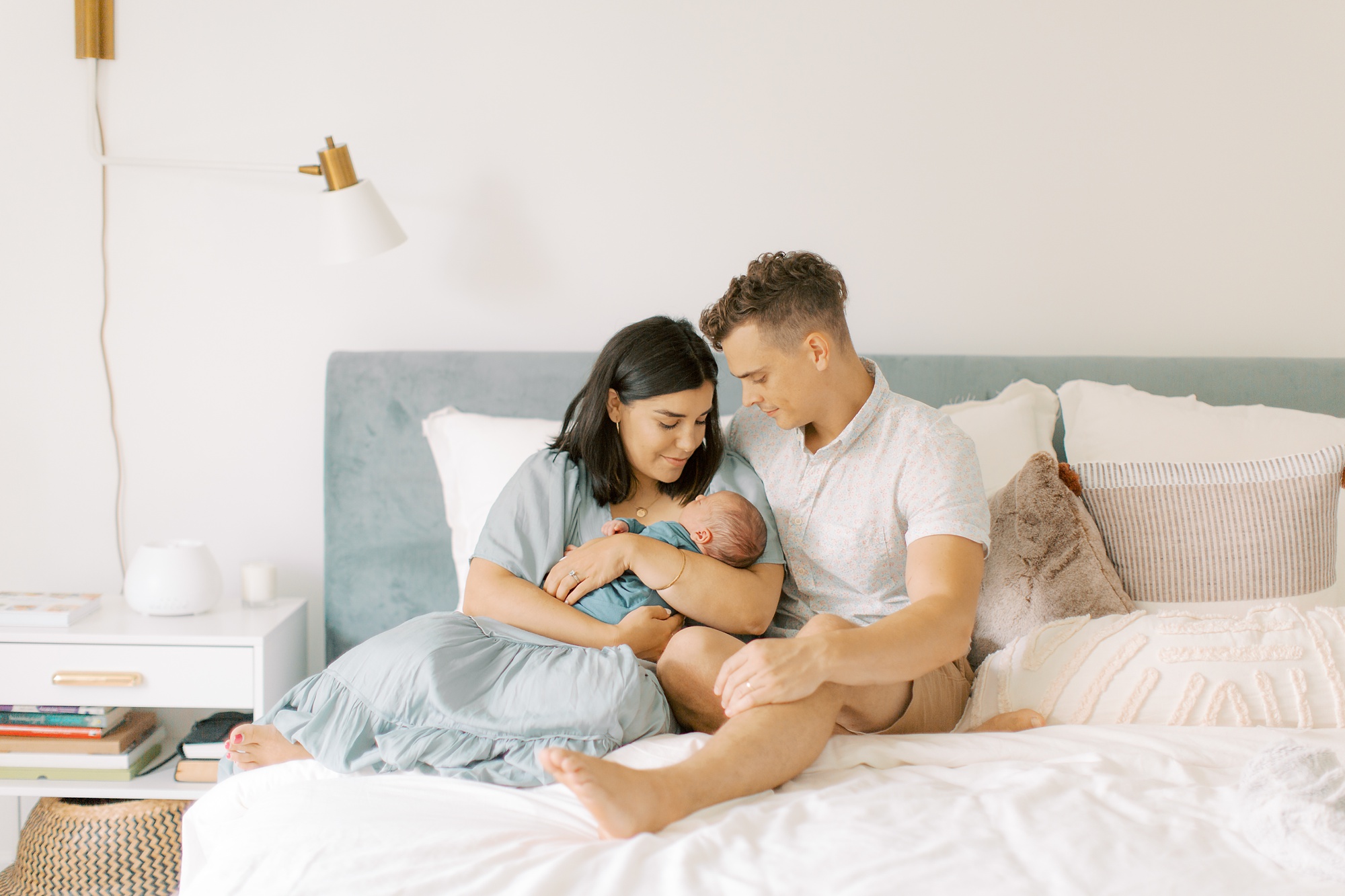 mom and dad sit on bed with white sheets holding new baby boy in blue wrap