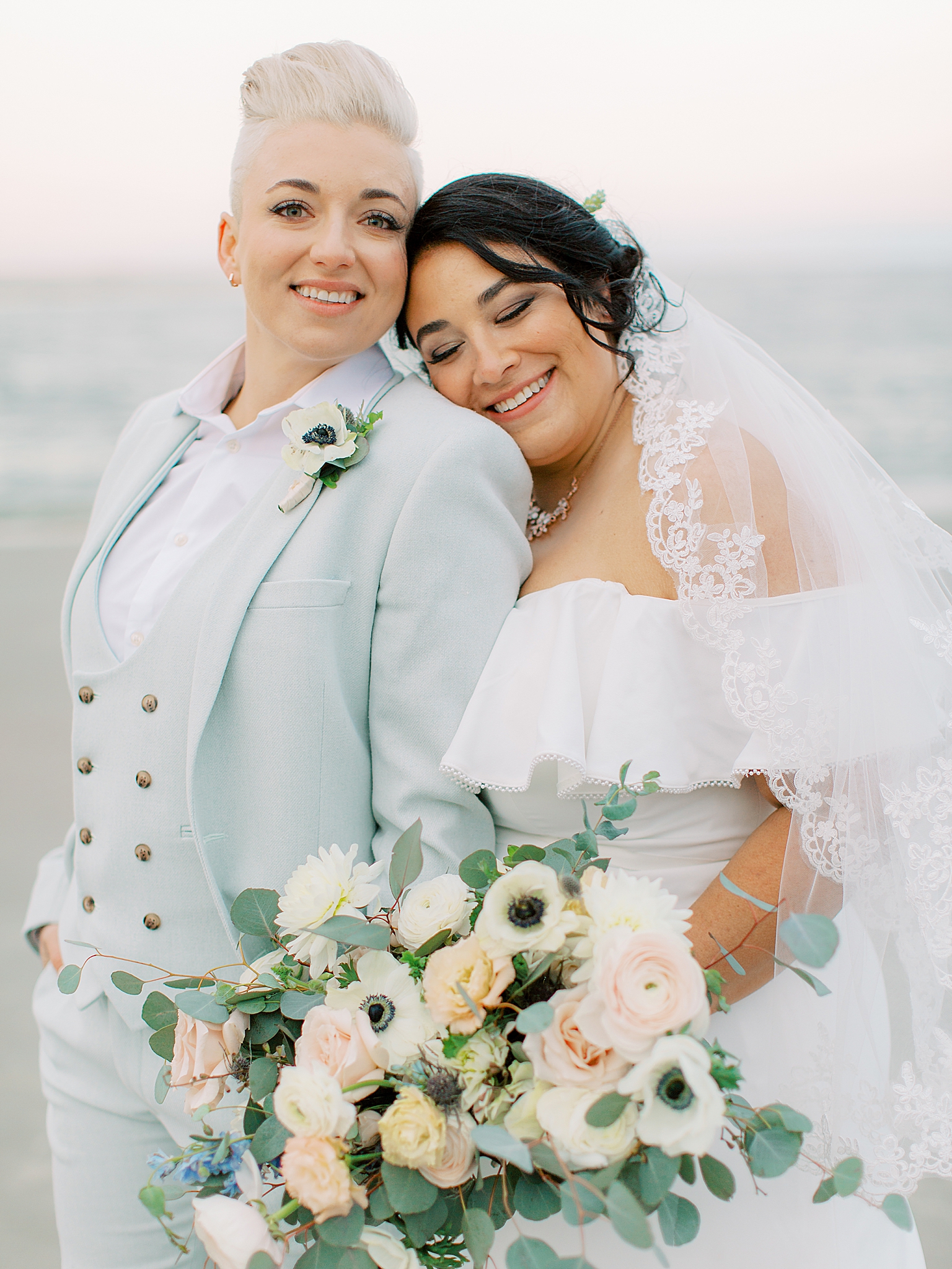 bride leans on partner's shoulder during photos on beach